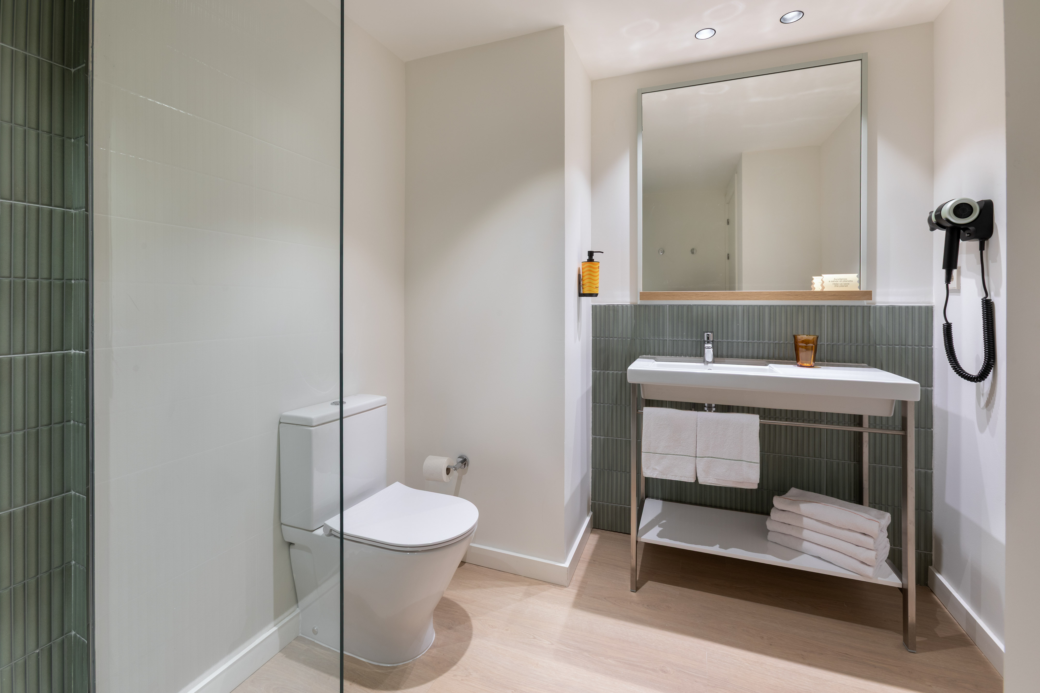 Modern bathroom with white toilet, sink, mirror, crisp towels, and green tiled wall.