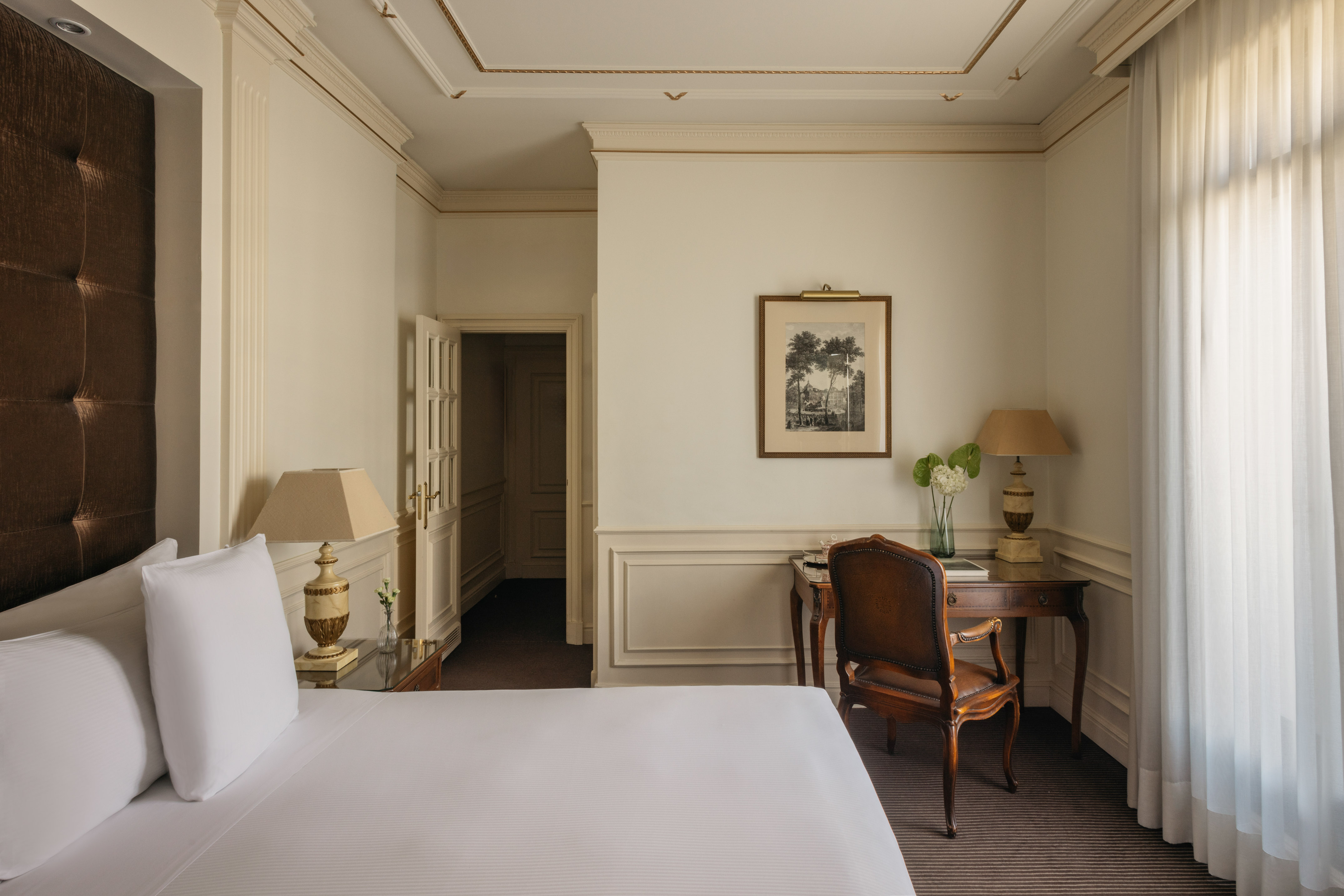 Luxurious hotel bedroom with white bed, classic wooden desk, and ornate molding.