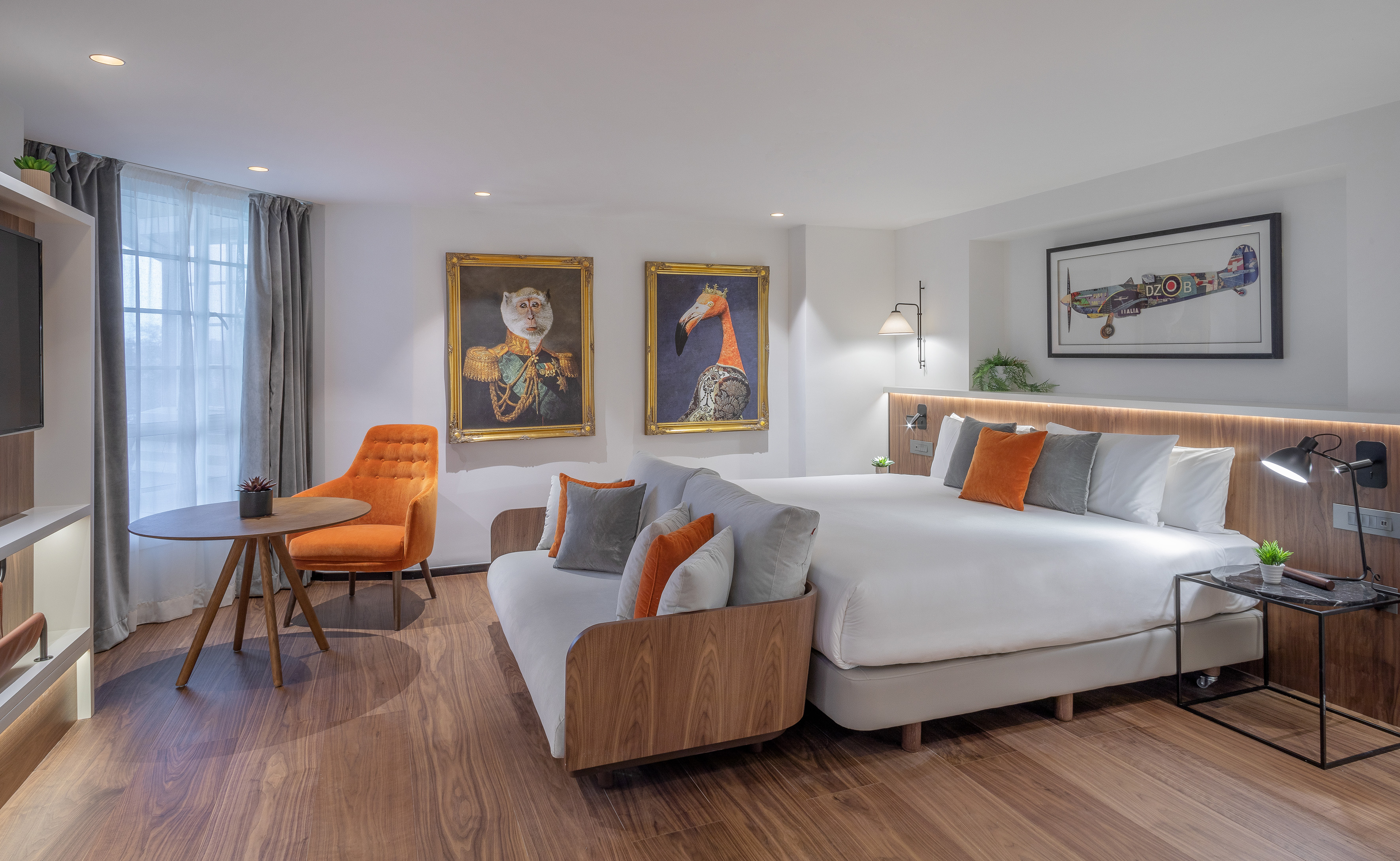 Contemporary hotel room: bed, sofa, orange chair, hardwood floor, decorative artwork.