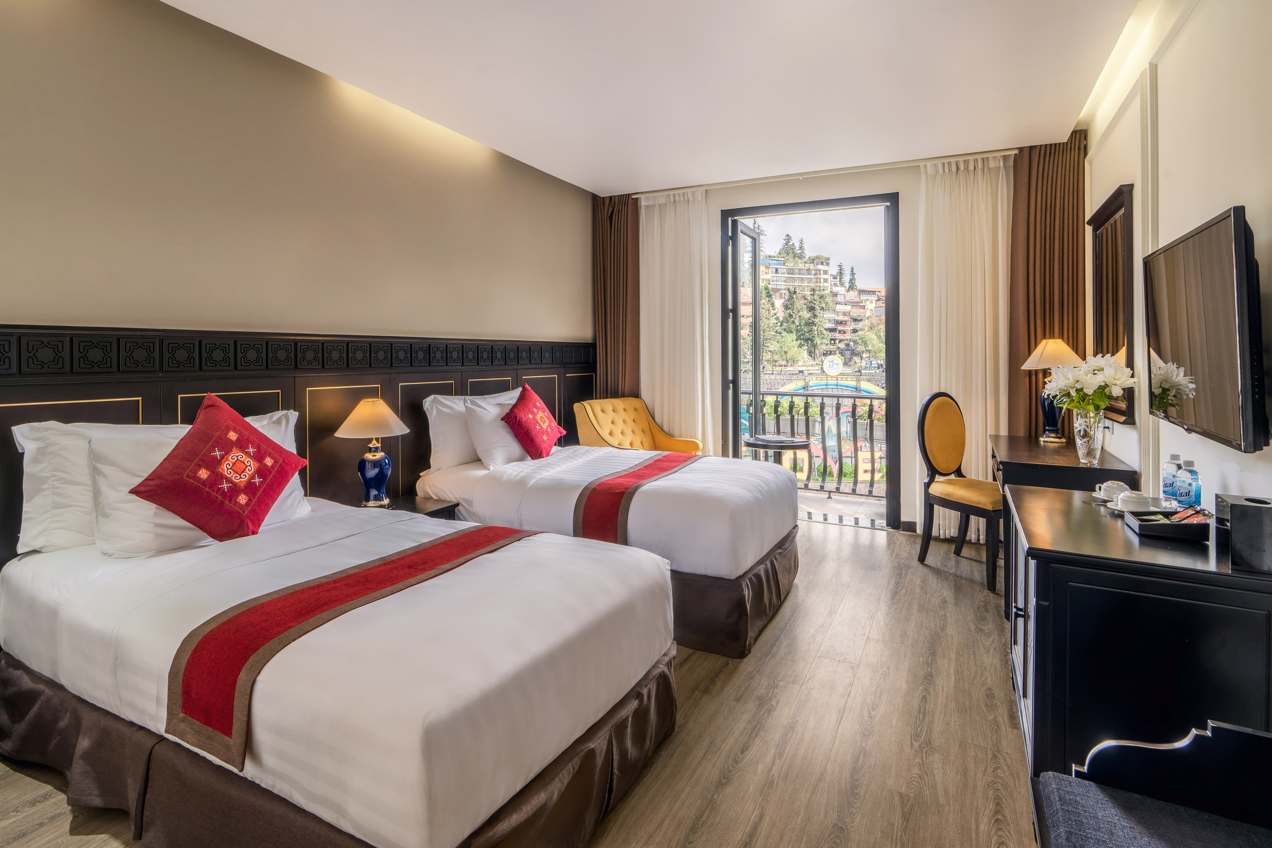 Hotel room with twin beds, red accents, and a balcony overlooking a town.