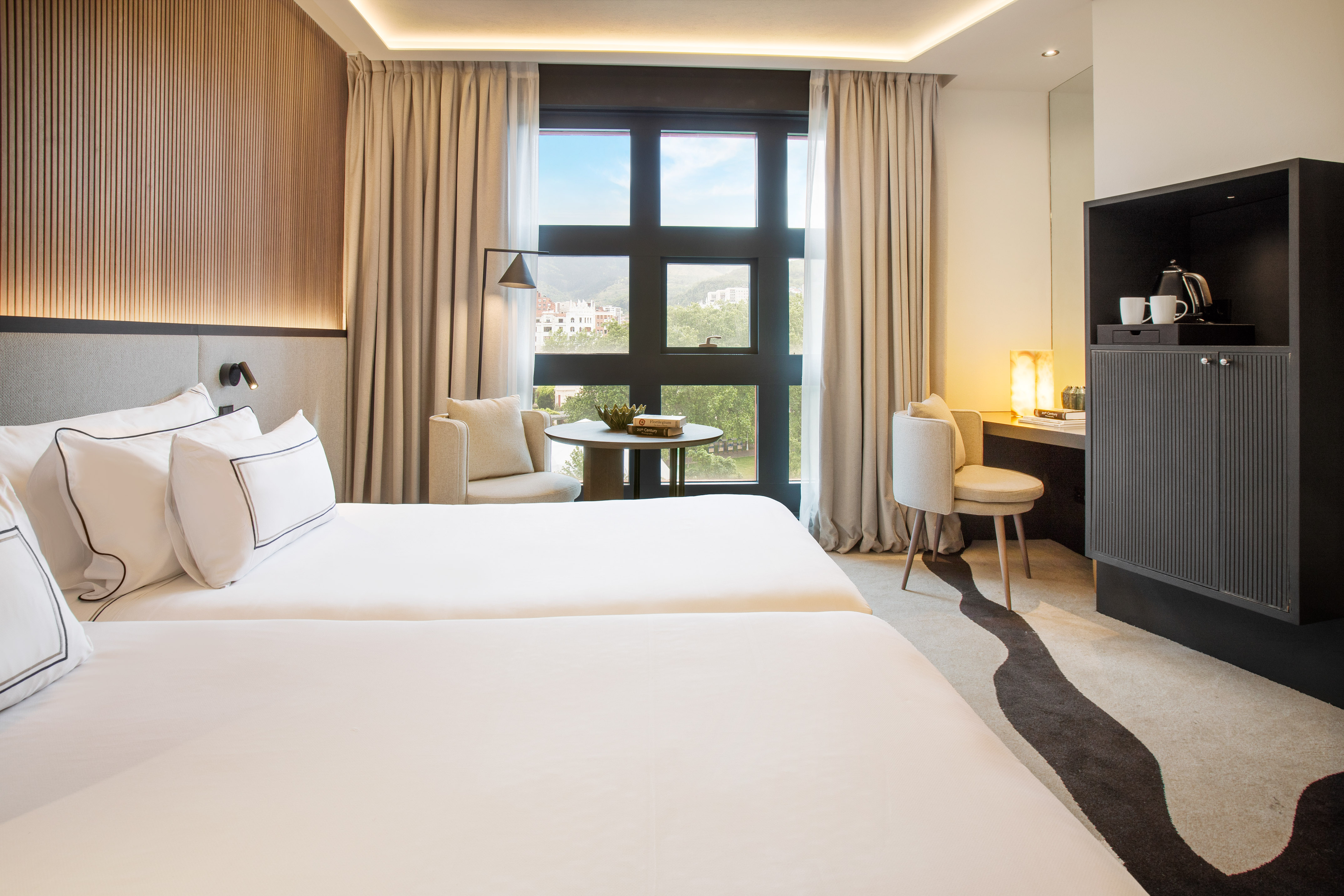 Modern twin hotel room with window view, desk, and coffee station.