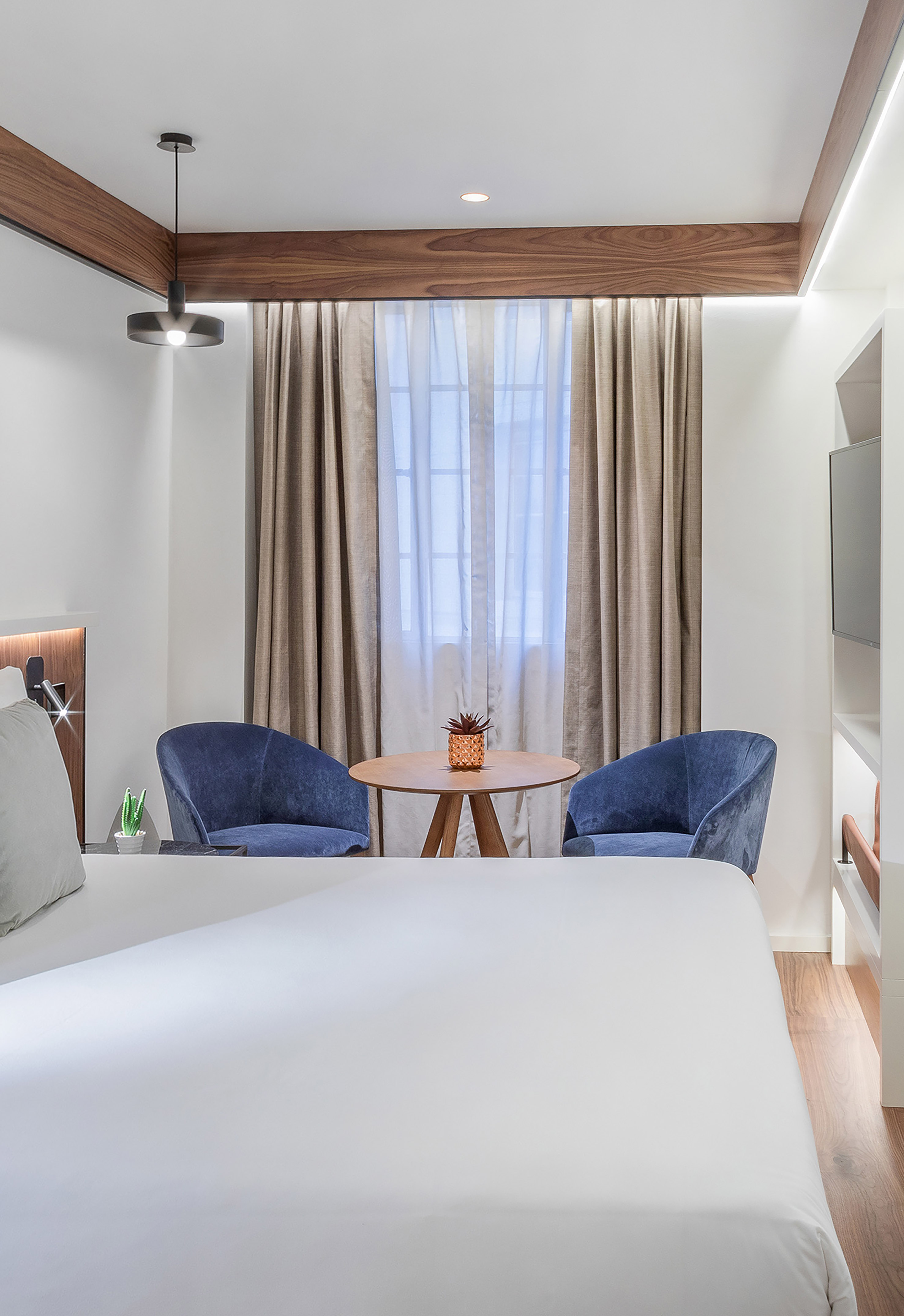 Modern hotel bedroom with white bed, blue armchairs, and wooden accents.