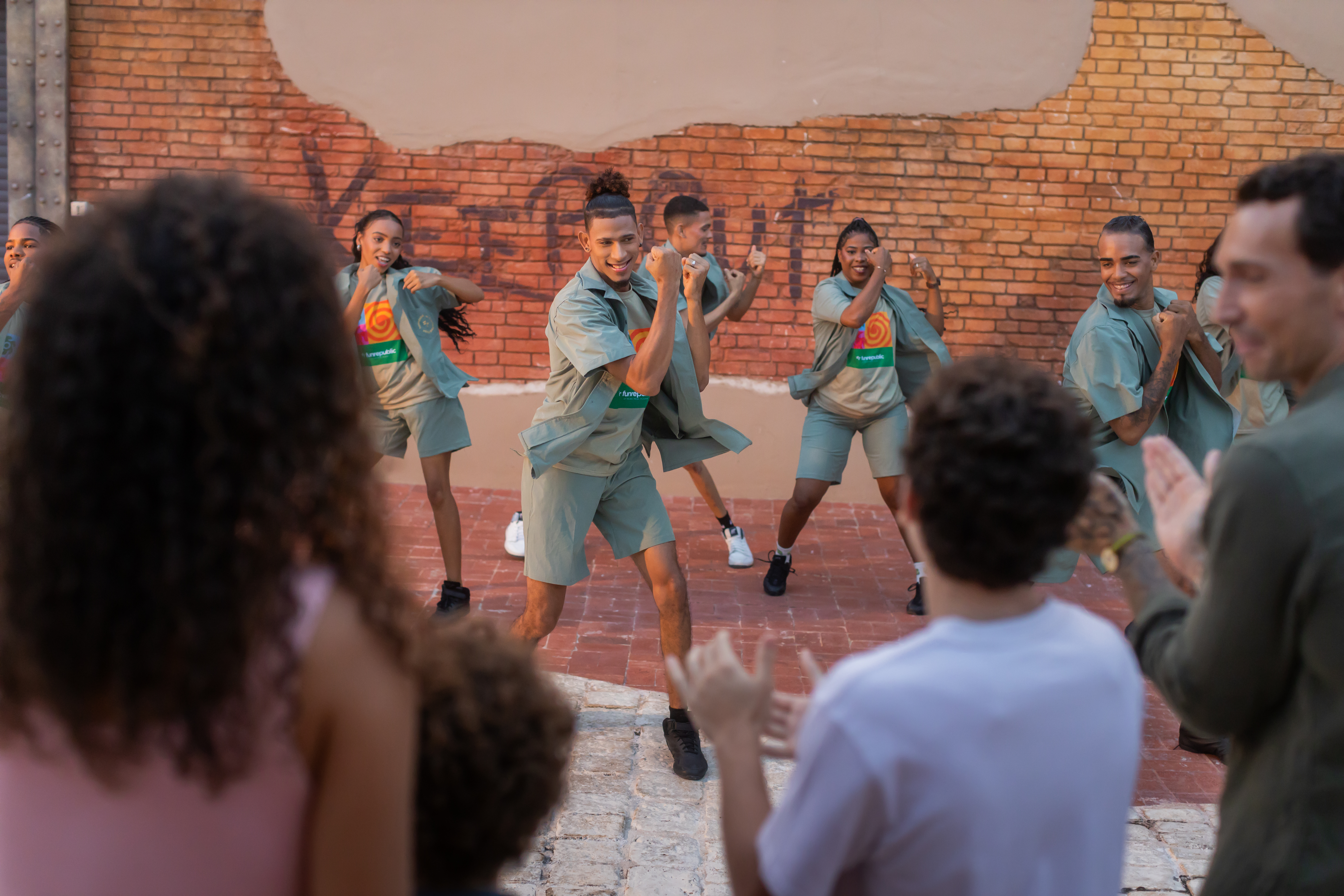 Energetic group dancing outdoors against a brick wall, with spectators clapping.