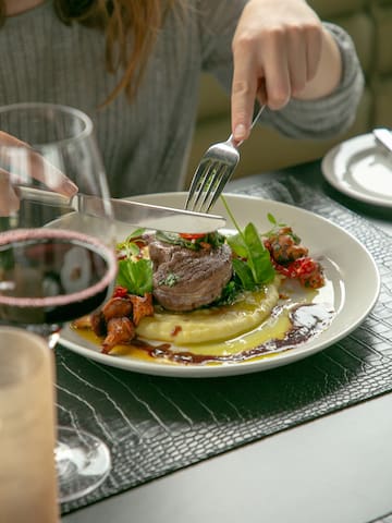 a person cutting a steak on a plate with a glass of wine