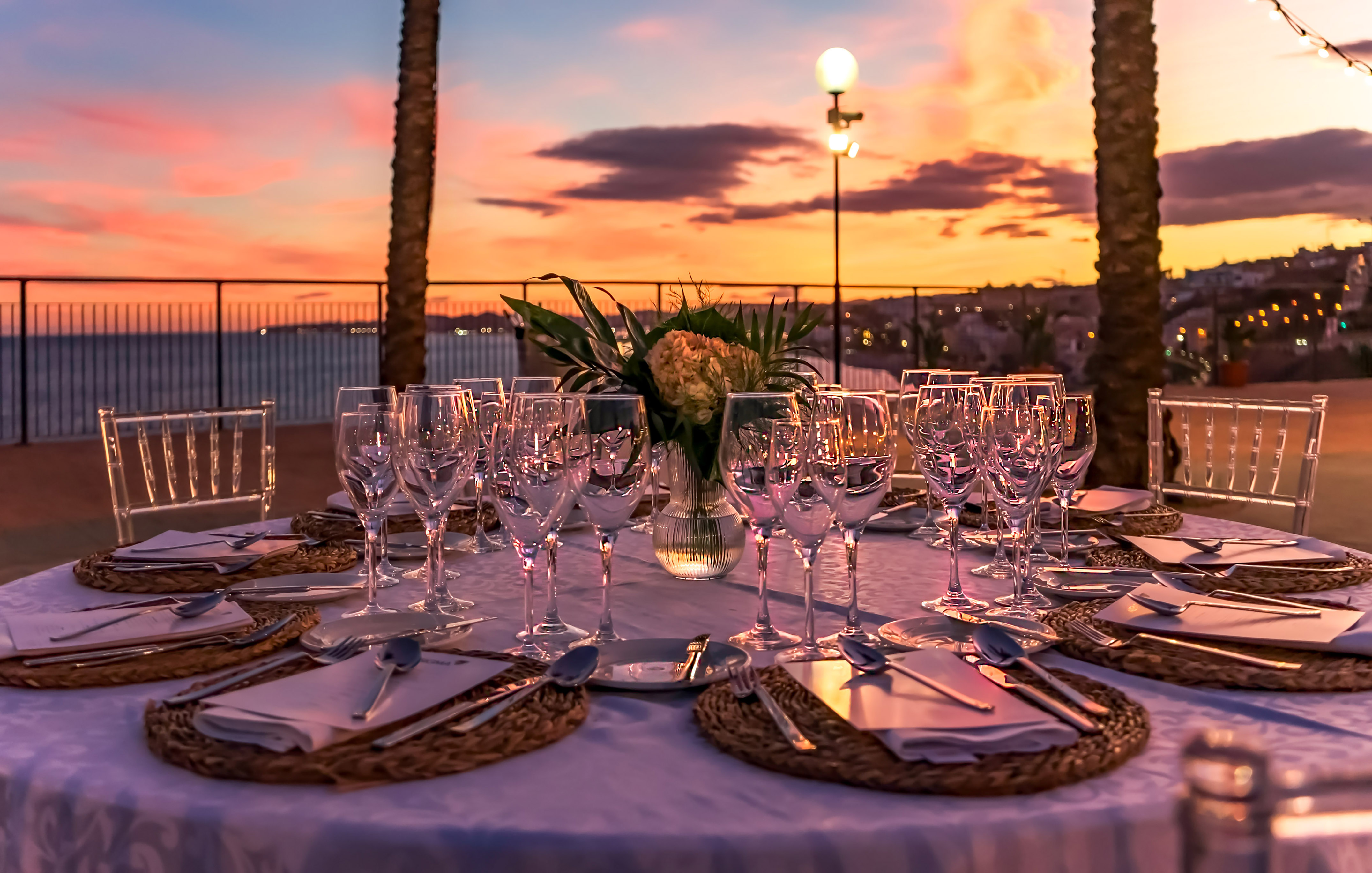 Outdoor elegant dining table setting with wine glasses and sunset ocean view.