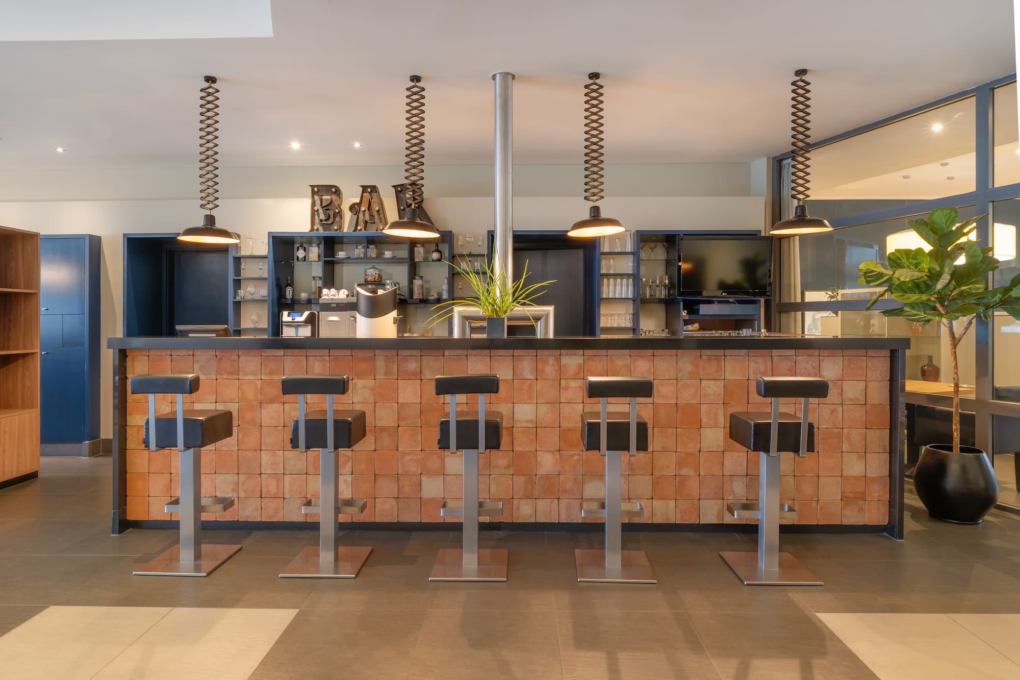 Modern bar counter with terracotta tiles, industrial lights, and black stools.