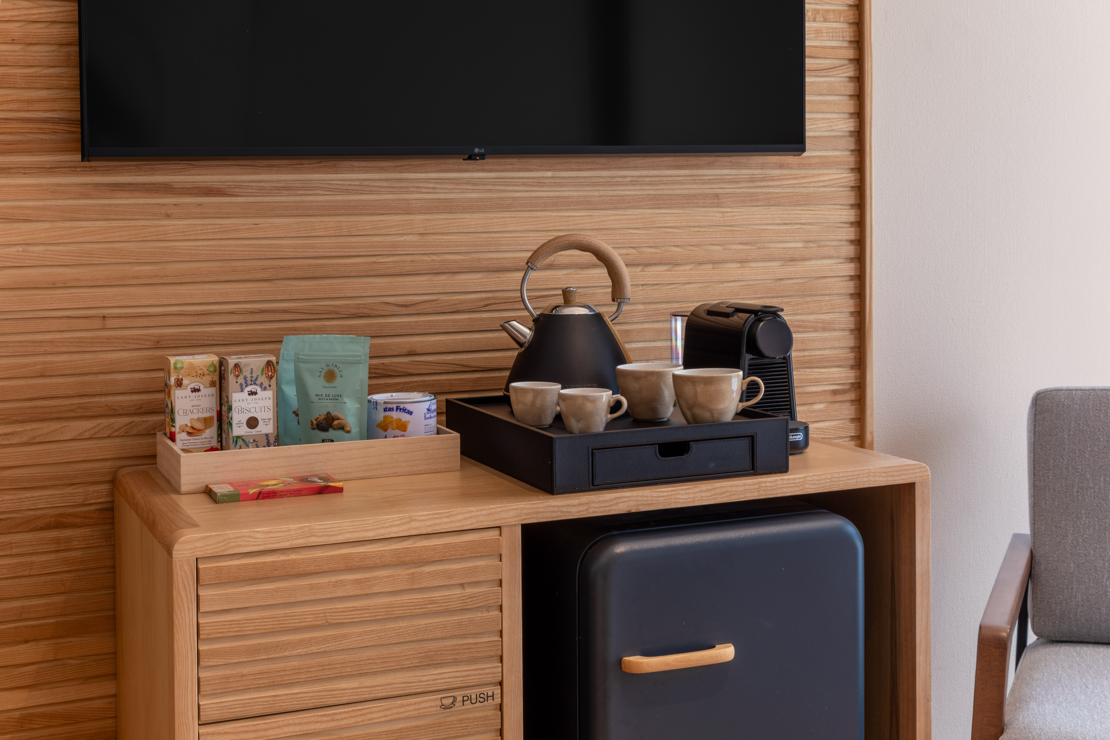 Mini bar de hotel: Nespresso, hervidor, tazas, snacks en mueble de madera.