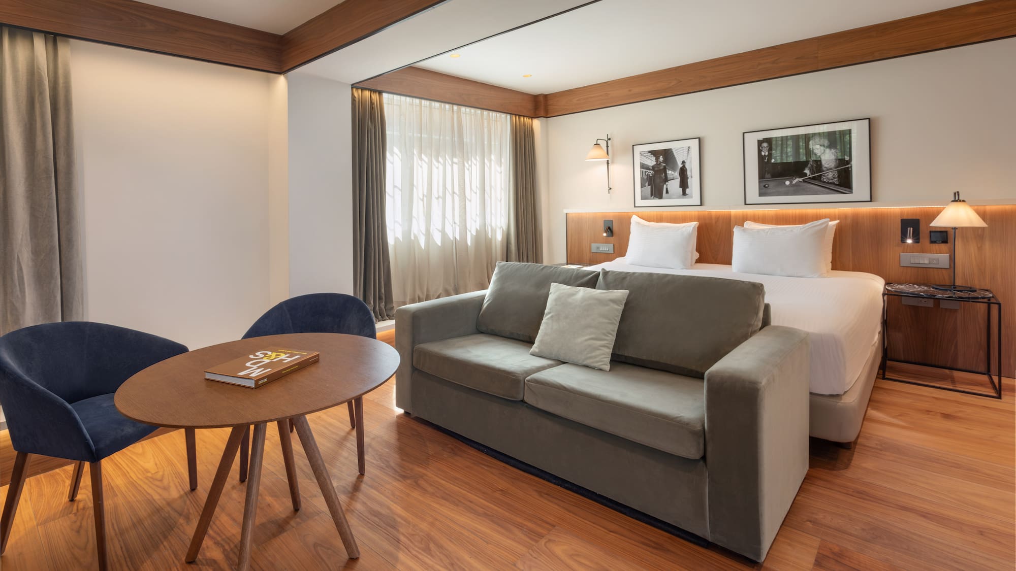 Modern hotel room with bed, grey sofa, wood floor, and seating area.