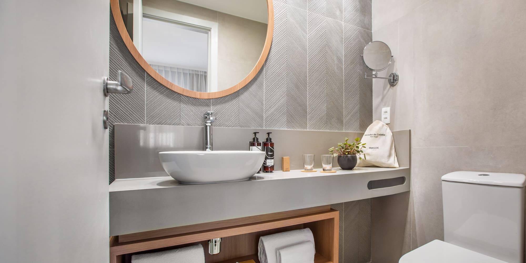 Modern hotel bathroom with round mirror, vessel sink, and toilet. Gray chevron tiles.