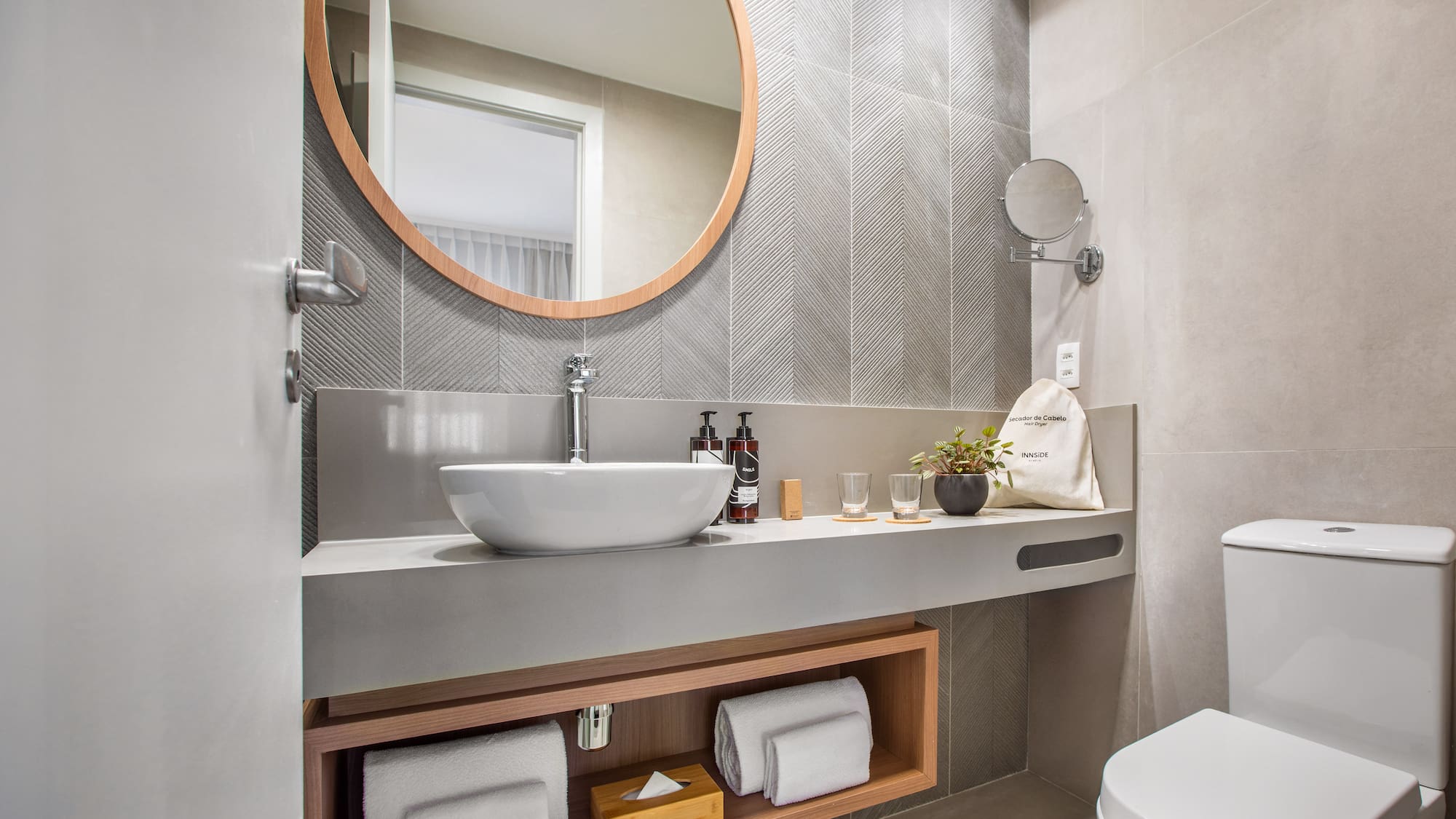 Modern hotel bathroom with round mirror, vessel sink, and toilet. Gray chevron tiles.
