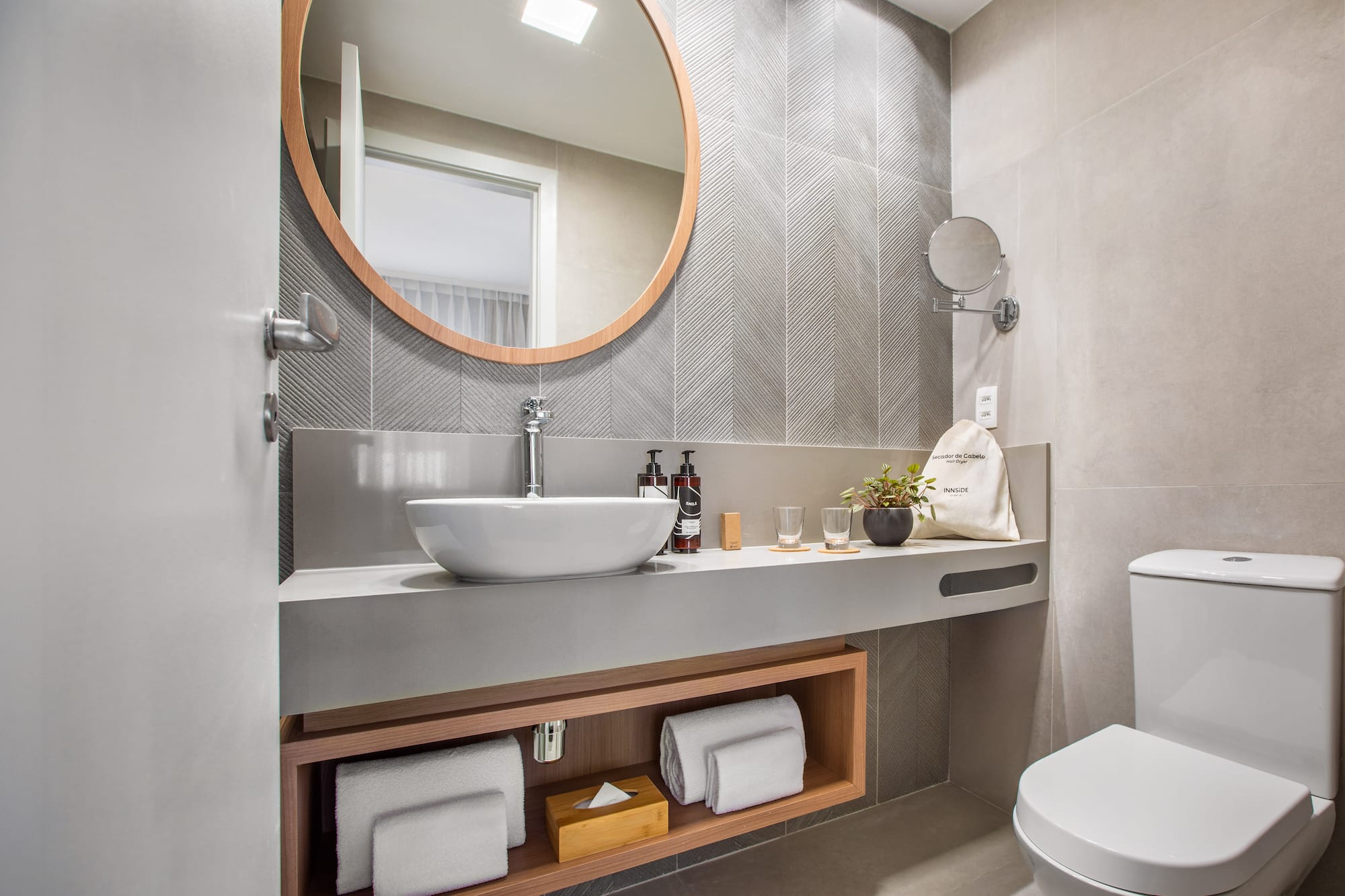 Modern hotel bathroom with round mirror, vessel sink, and toilet. Gray chevron tiles.