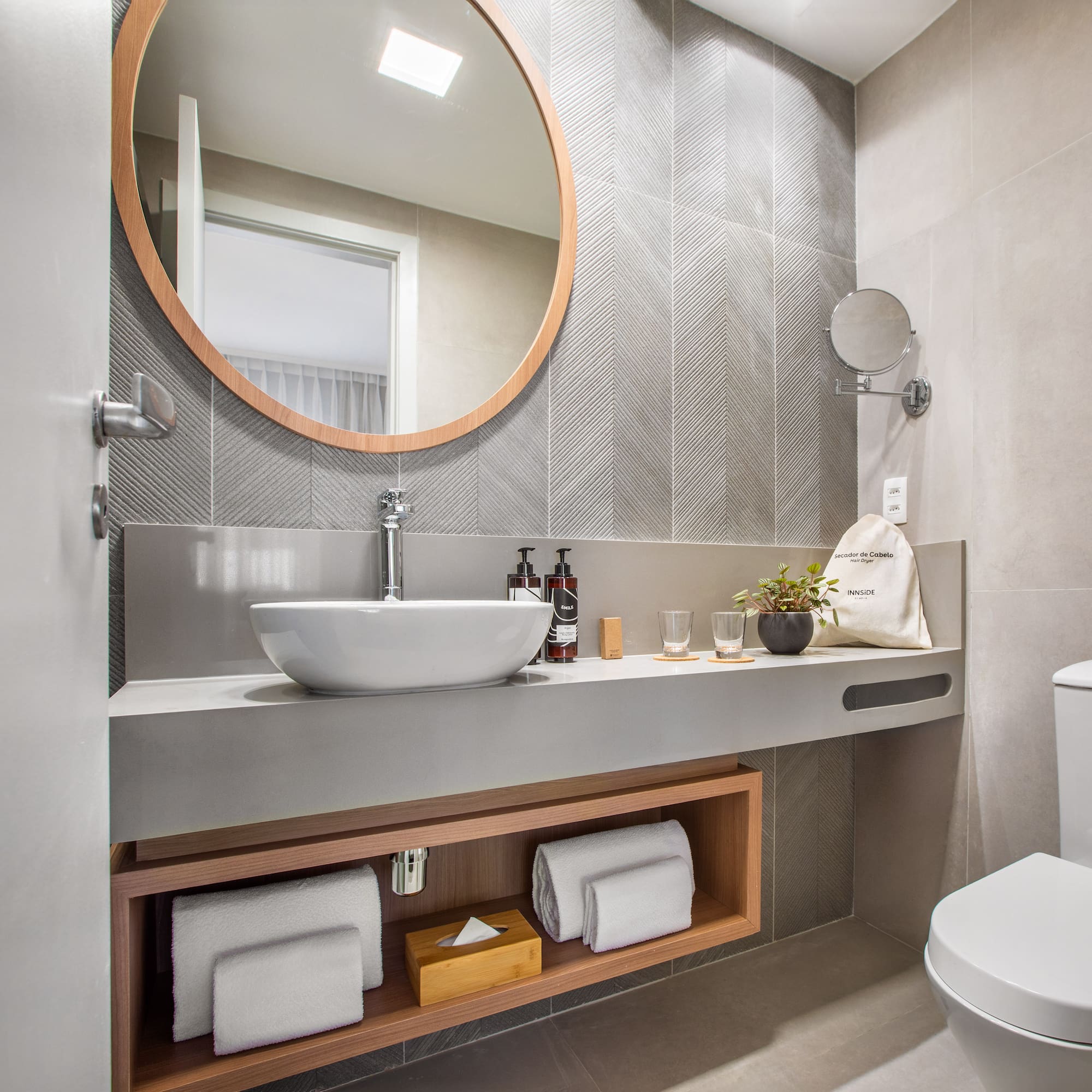 Modern hotel bathroom with round mirror, vessel sink, and toilet. Gray chevron tiles.