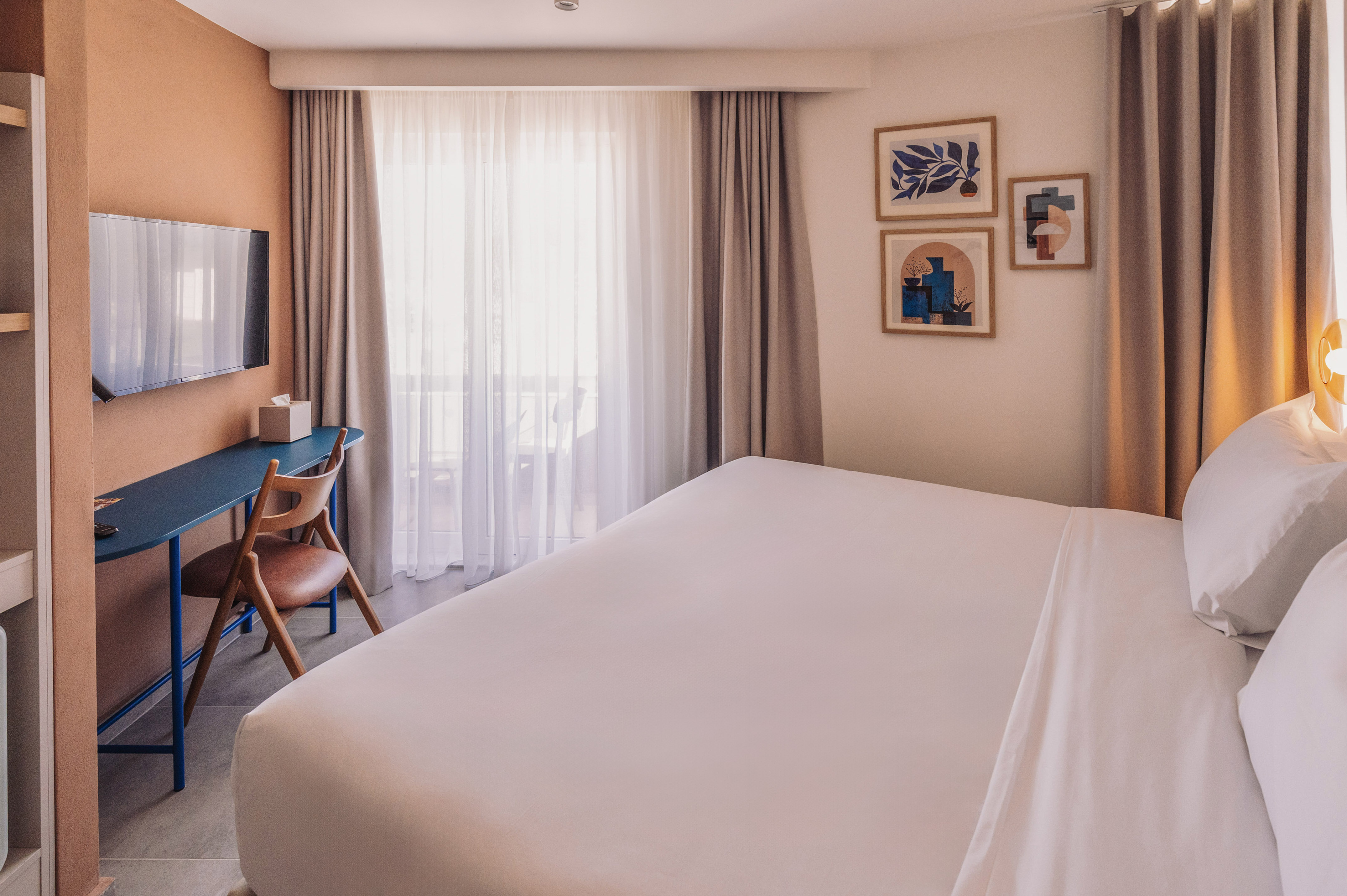Bright modern hotel room with king bed, blue desk, TV, and abstract art.