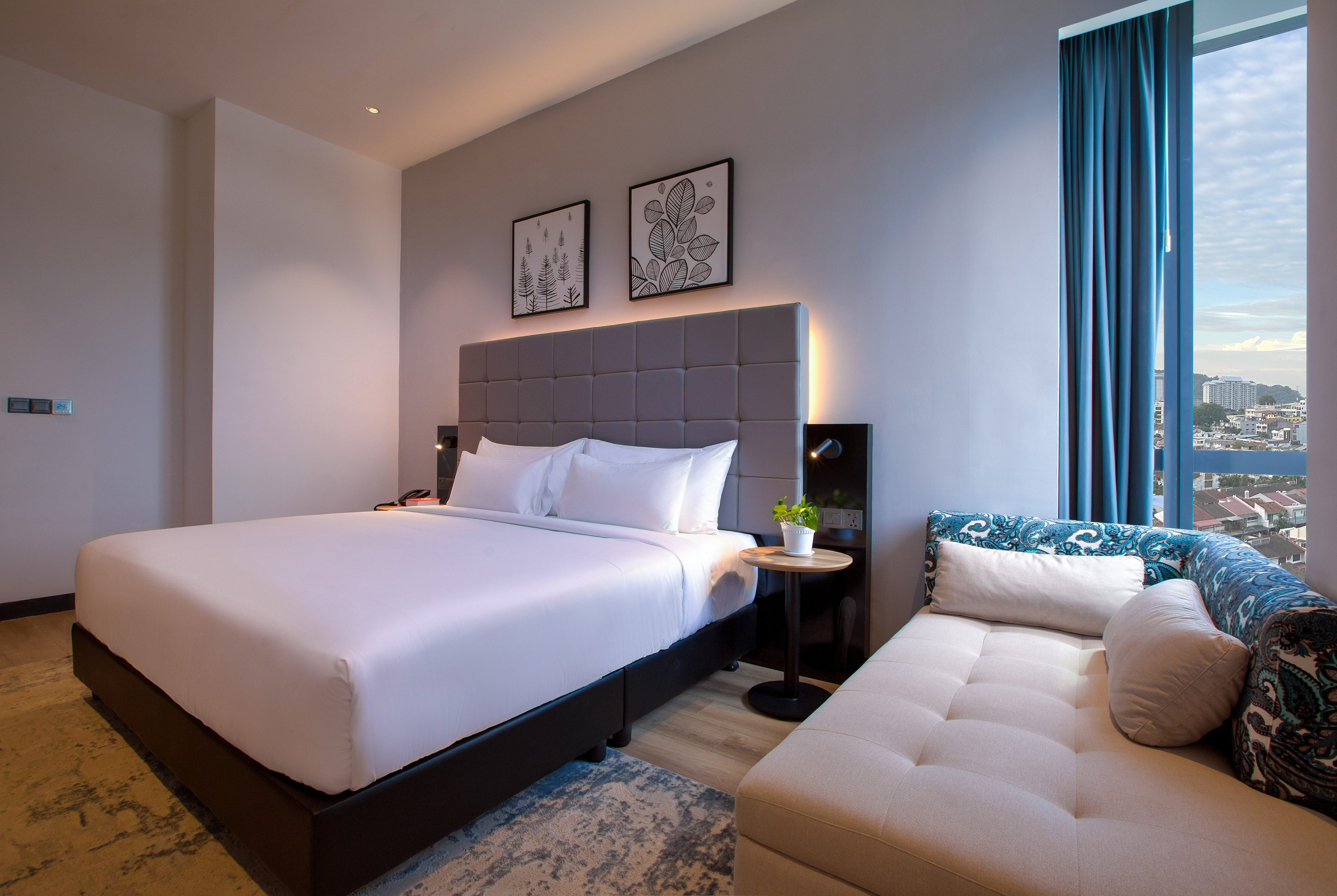Modern hotel bedroom with king bed, chaise lounge, and city skyline view.