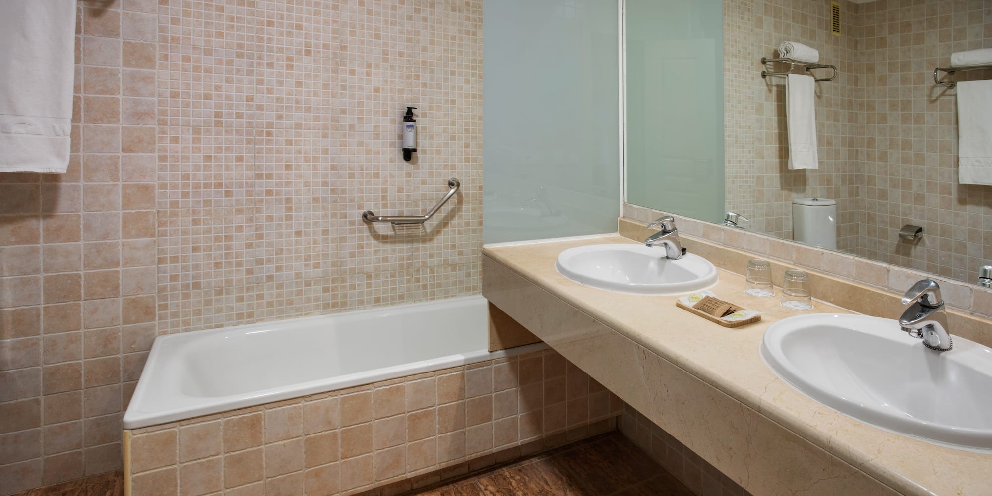 Hotel bathroom with bathtub, double vanity, mirror, and mosaic tiles.