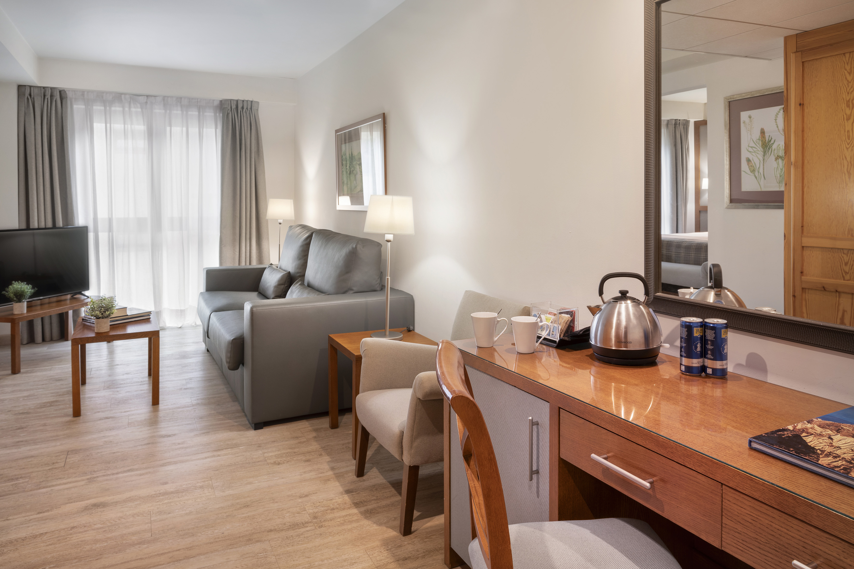Modern hotel suite living room with grey sofa, desk, TV, and wooden floor.