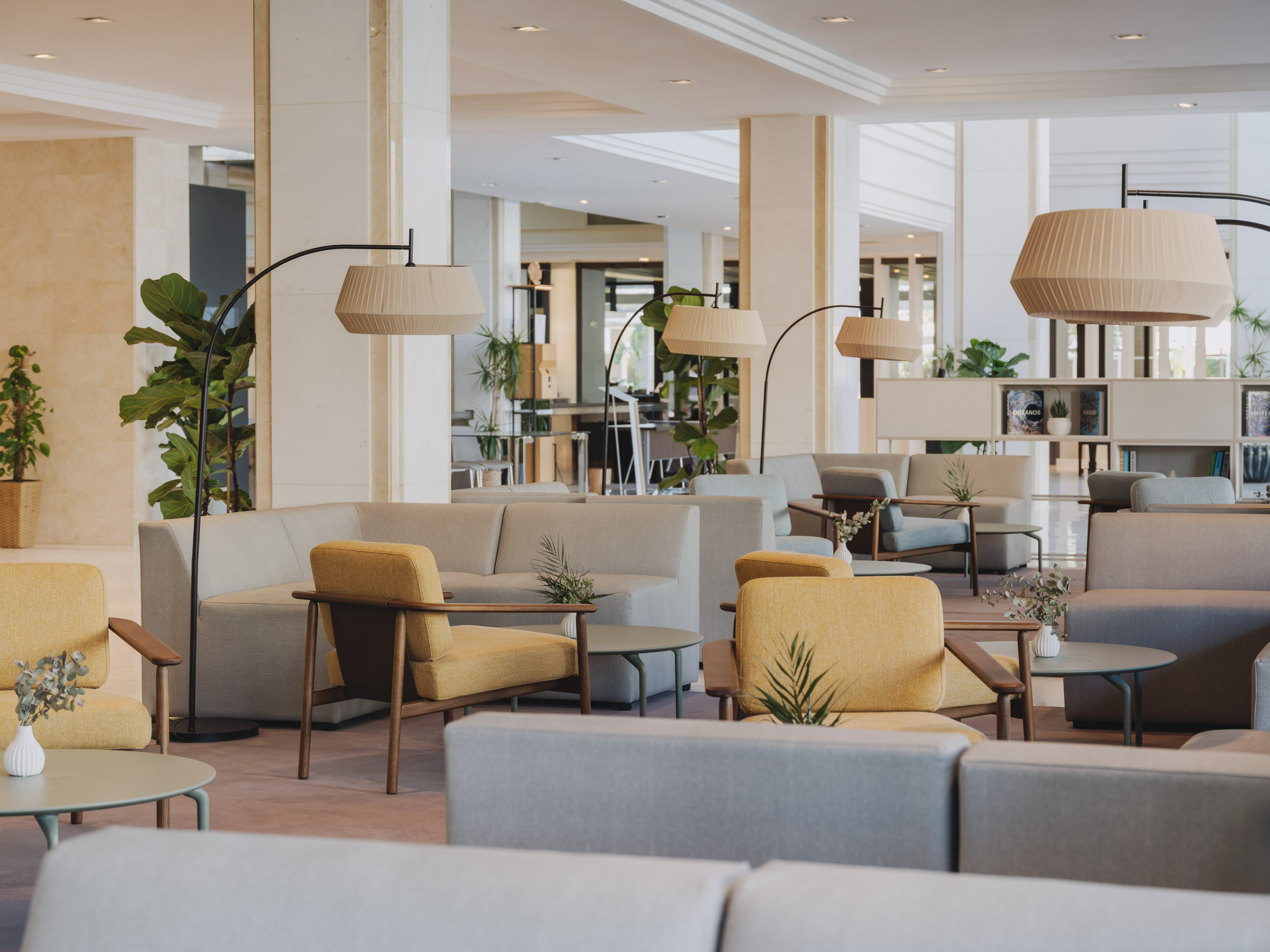 Modern hotel lounge with gray sofas, yellow armchairs, arc lamps, and green plants.
