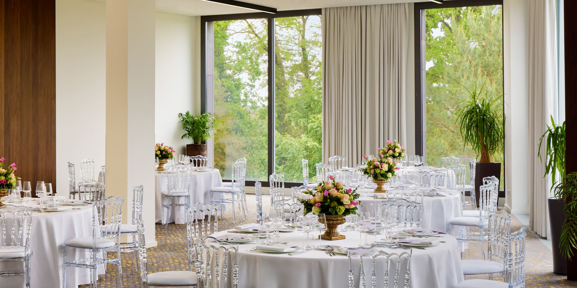 Elegant event space with set tables, clear chairs, flowers, and garden view.