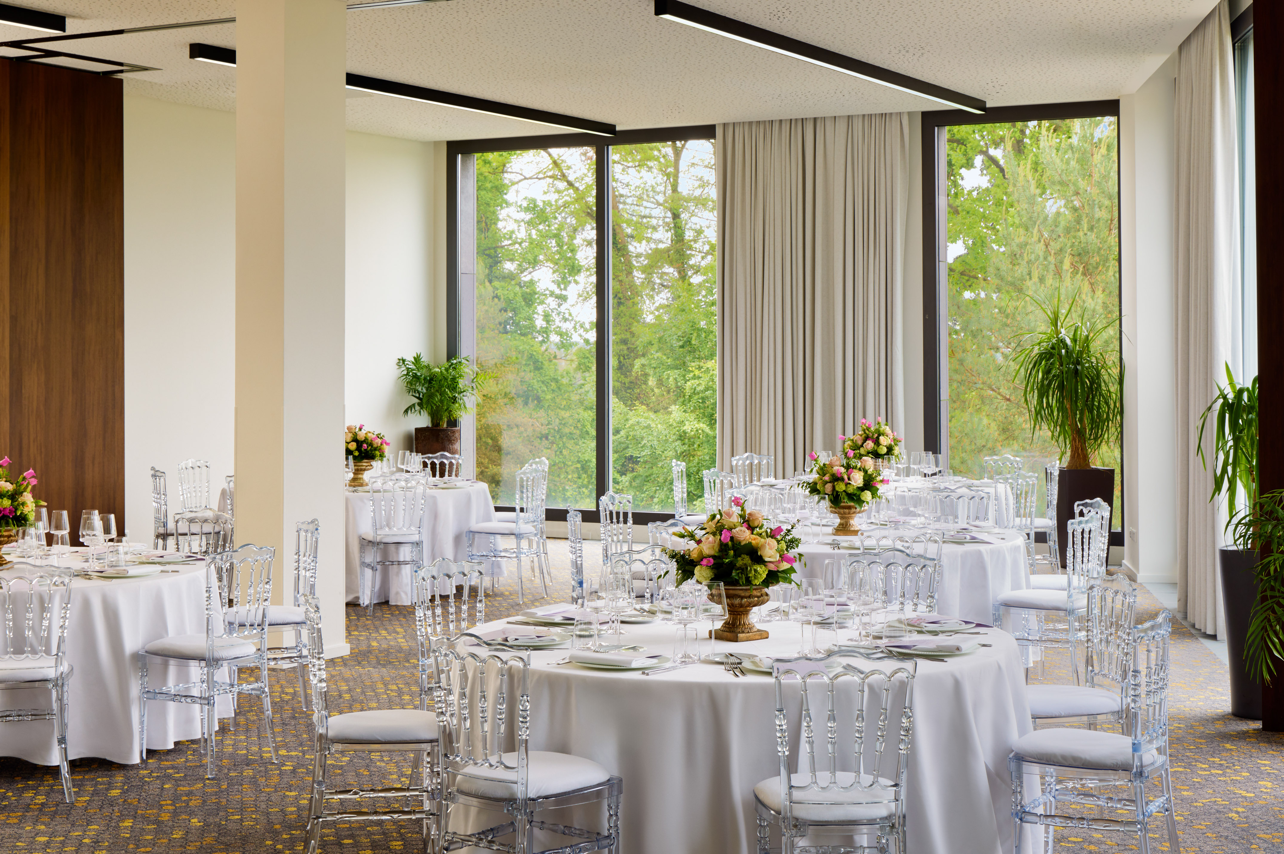 Elegant event space with set tables, clear chairs, flowers, and garden view.