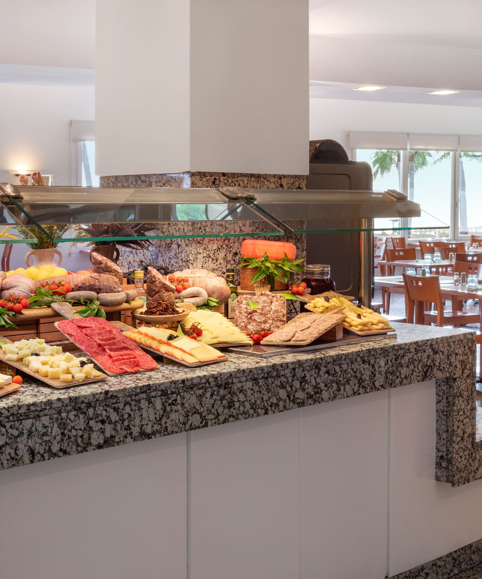 Hotel buffet with meats, cheeses, and fruits in a brightly lit dining room.