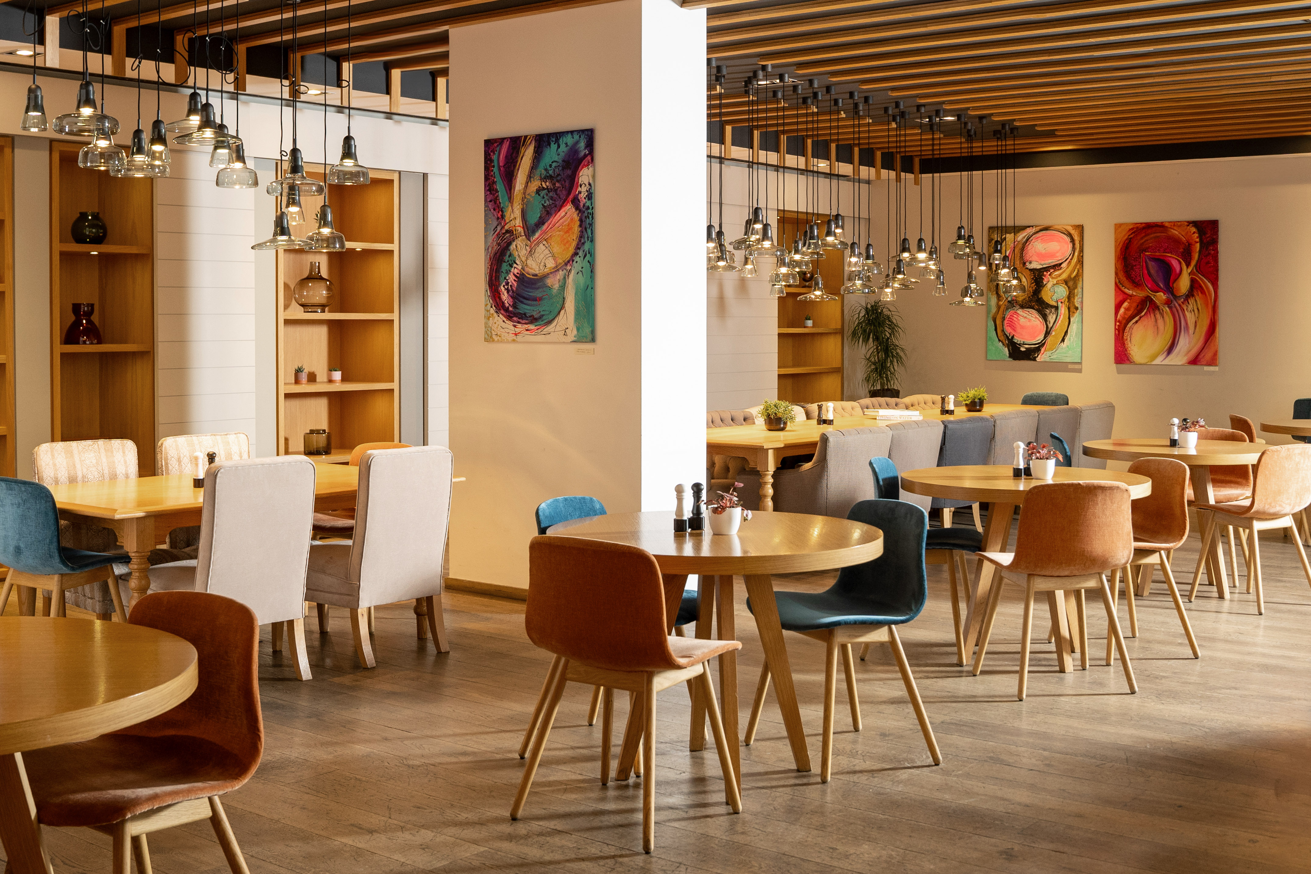 Modern restaurant dining room with wooden tables, varied chairs, abstract art, and pendant lights.