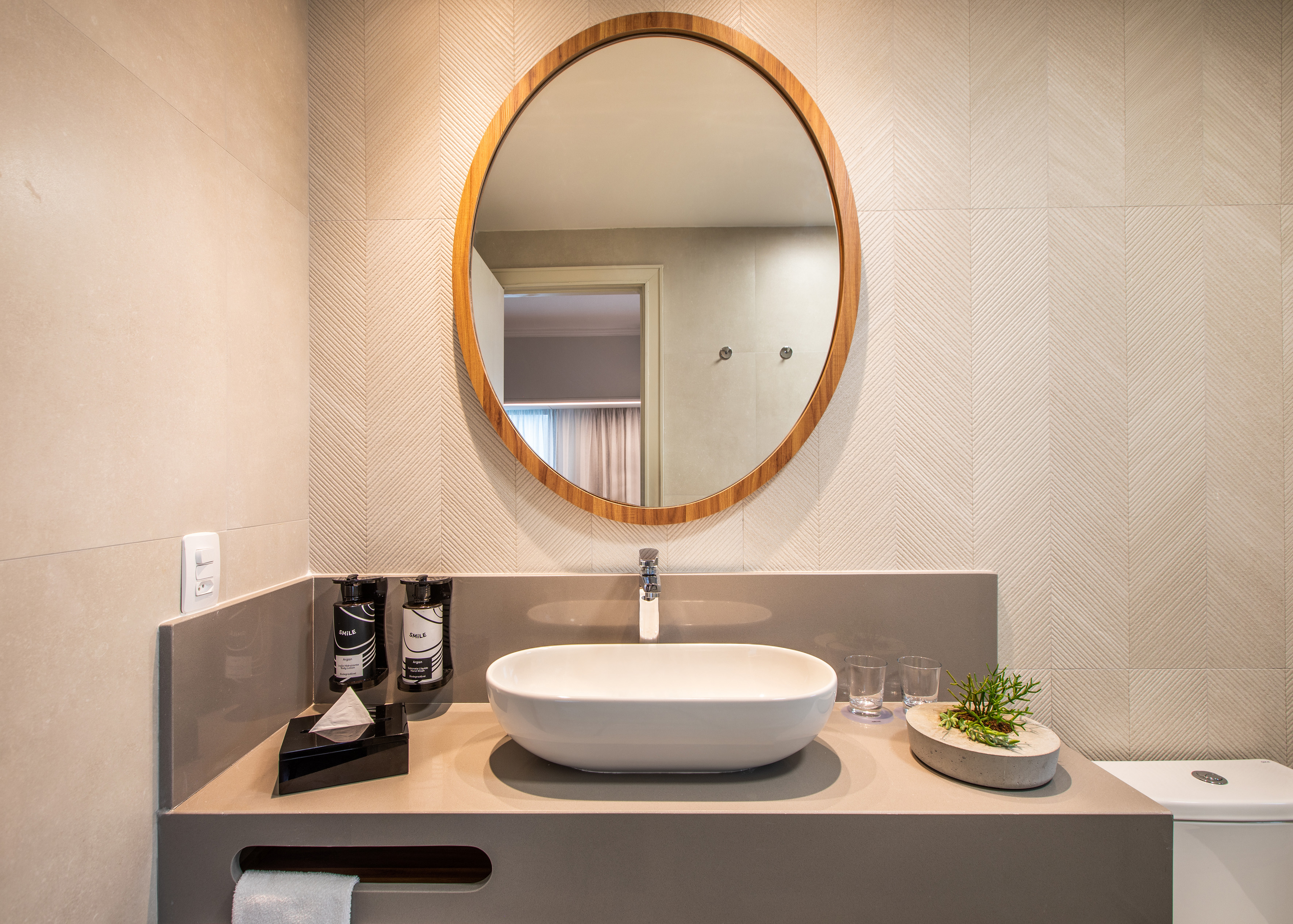 Modern hotel bathroom with oval mirror, vessel sink, and textured wall tiles.