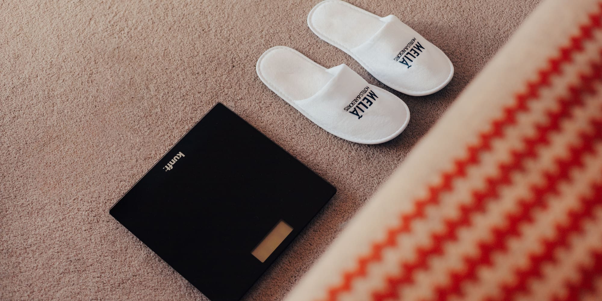 White Meliá hotel slippers and digital bathroom scale on carpet.