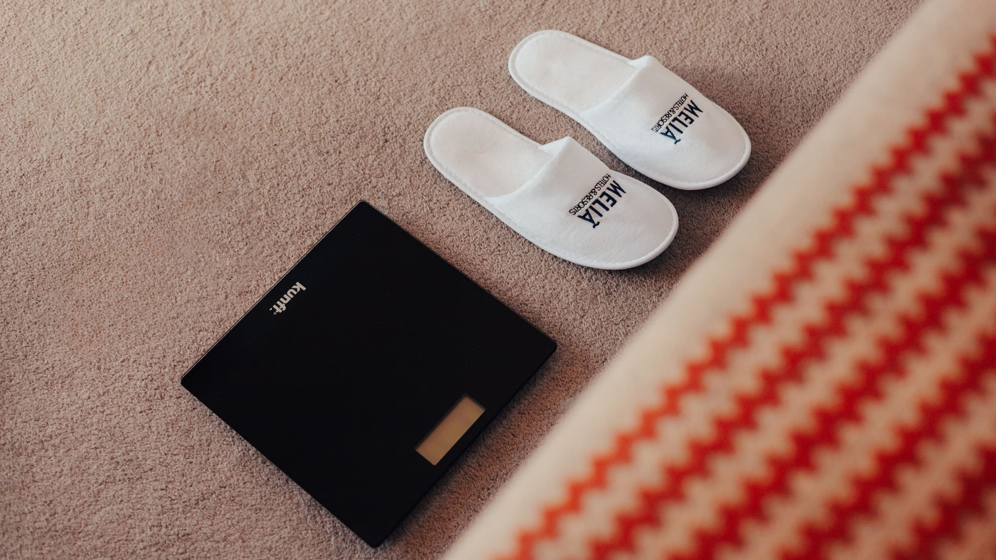 White Meliá hotel slippers and digital bathroom scale on carpet.