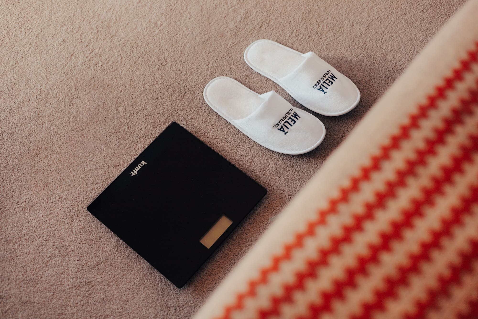 White Meliá hotel slippers and digital bathroom scale on carpet.