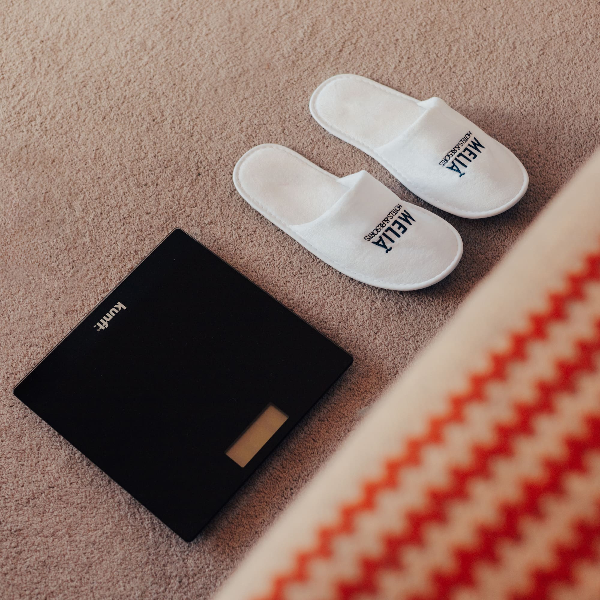 White Meliá hotel slippers and digital bathroom scale on carpet.
