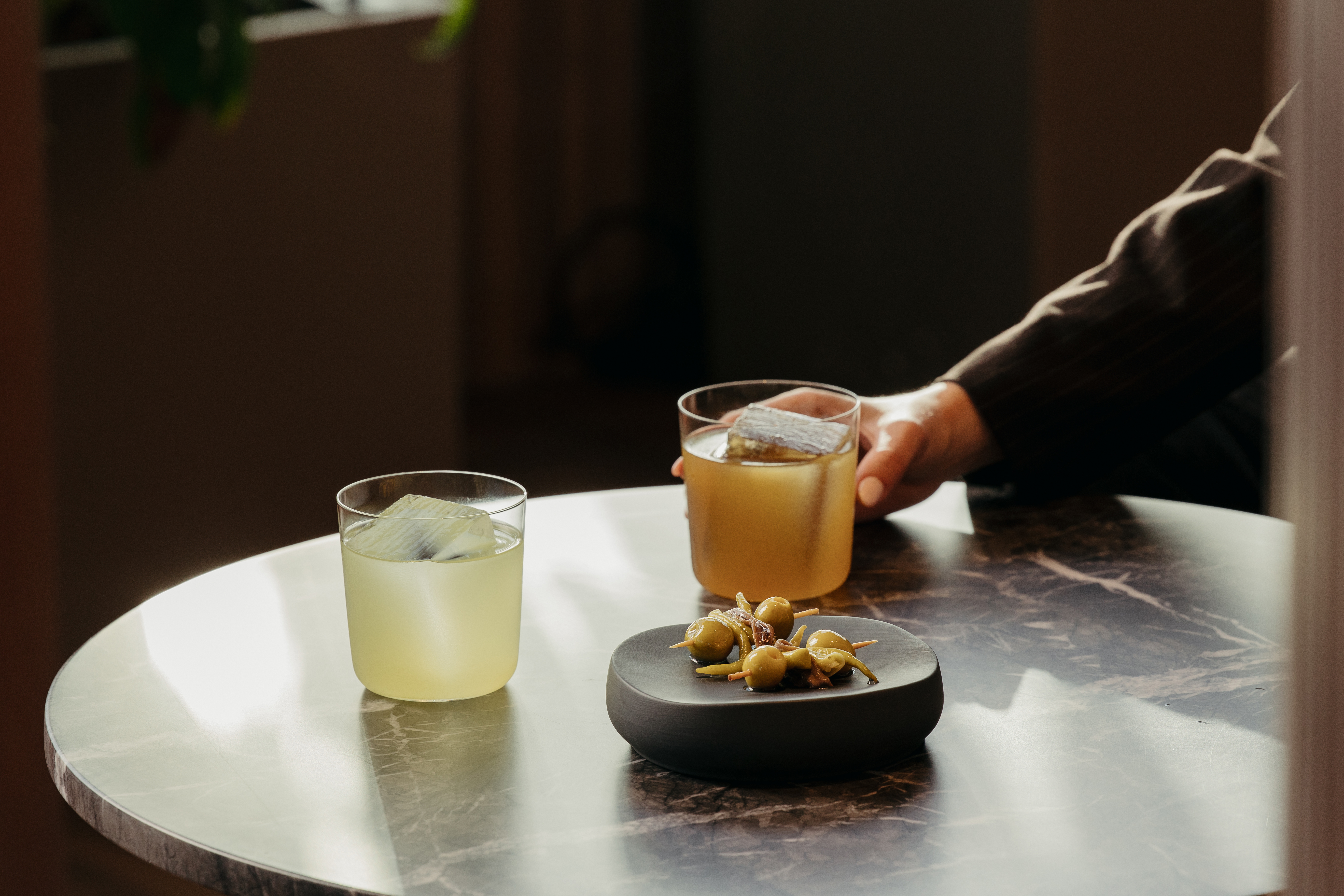 Two cocktails with ice, olives on a plate, and a hand on a marble table.