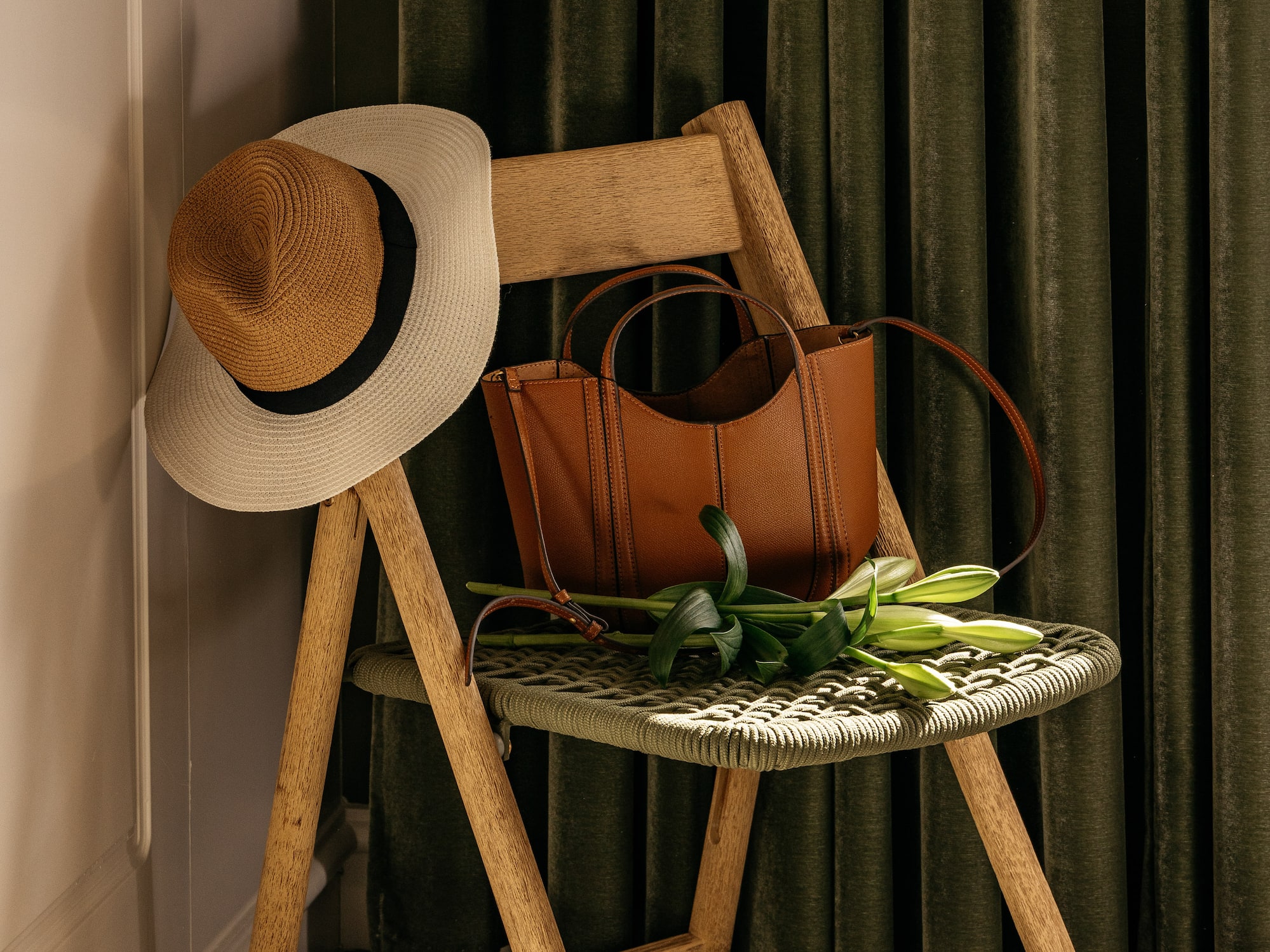 Silla madera plegable con sombrero paja, bolso marrón y lirios. Cortinas verdes.