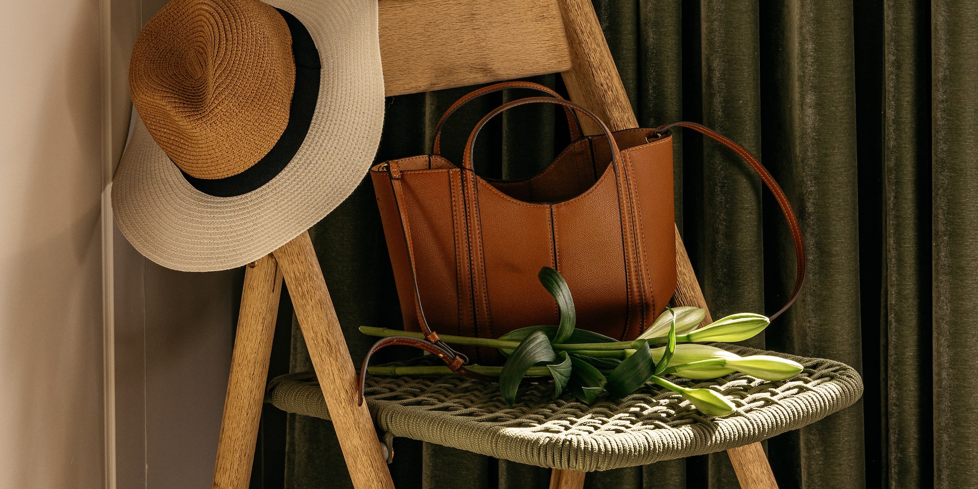 Silla madera plegable con sombrero paja, bolso marrón y lirios. Cortinas verdes.
