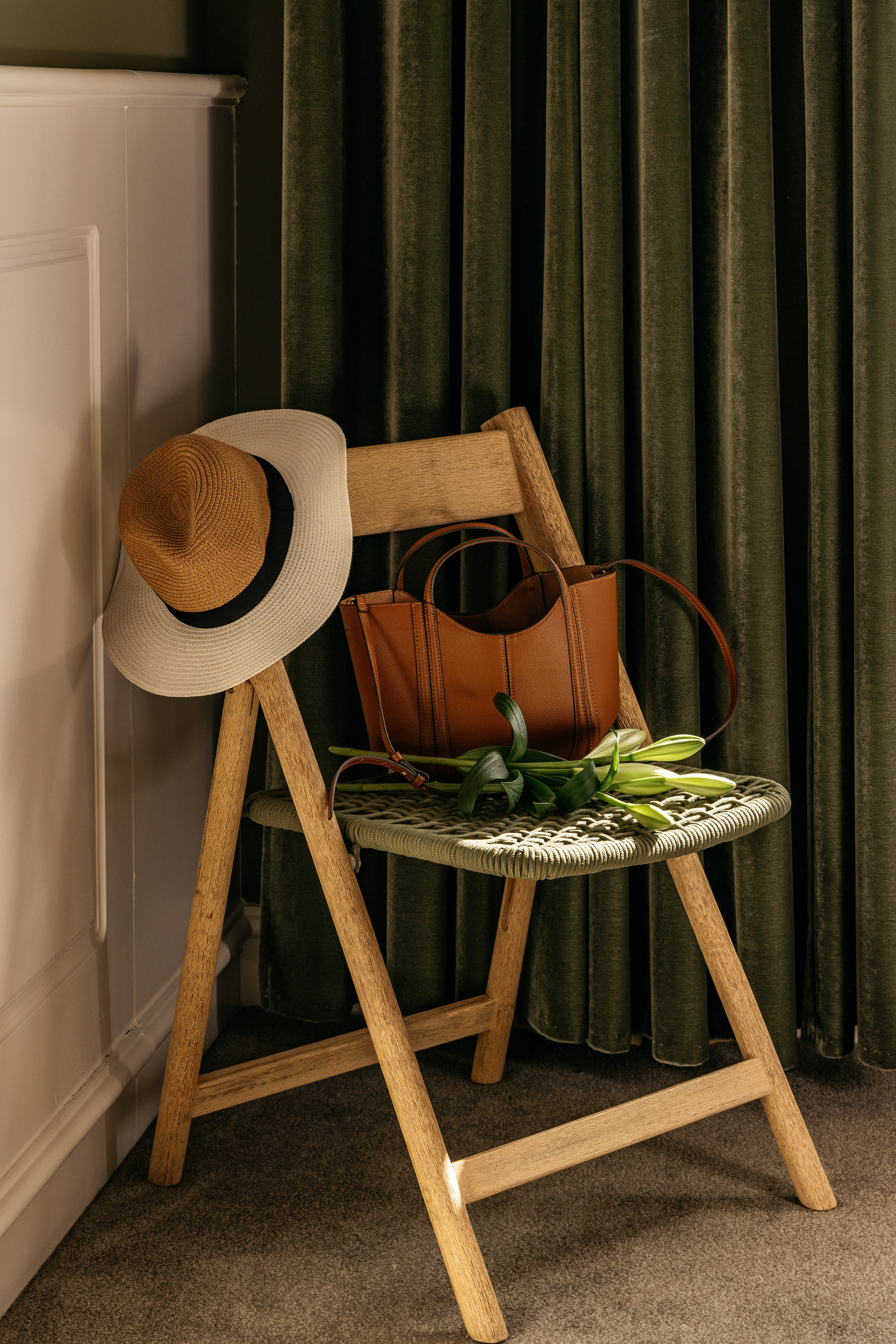 Silla madera plegable con sombrero paja, bolso marrón y lirios. Cortinas verdes.