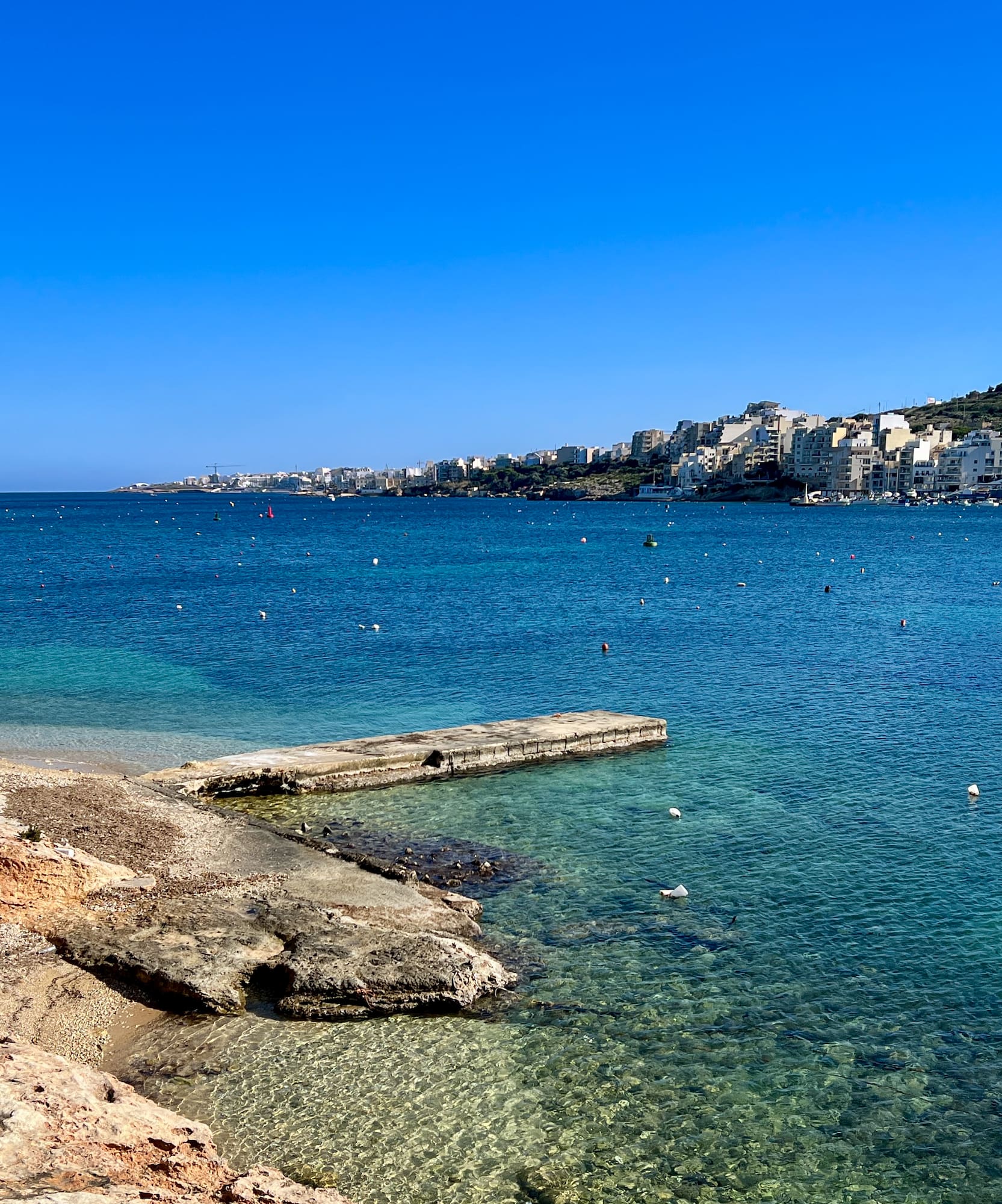 Malta: Bahía azul con pueblo costero, muelle y playa rocosa con bote.