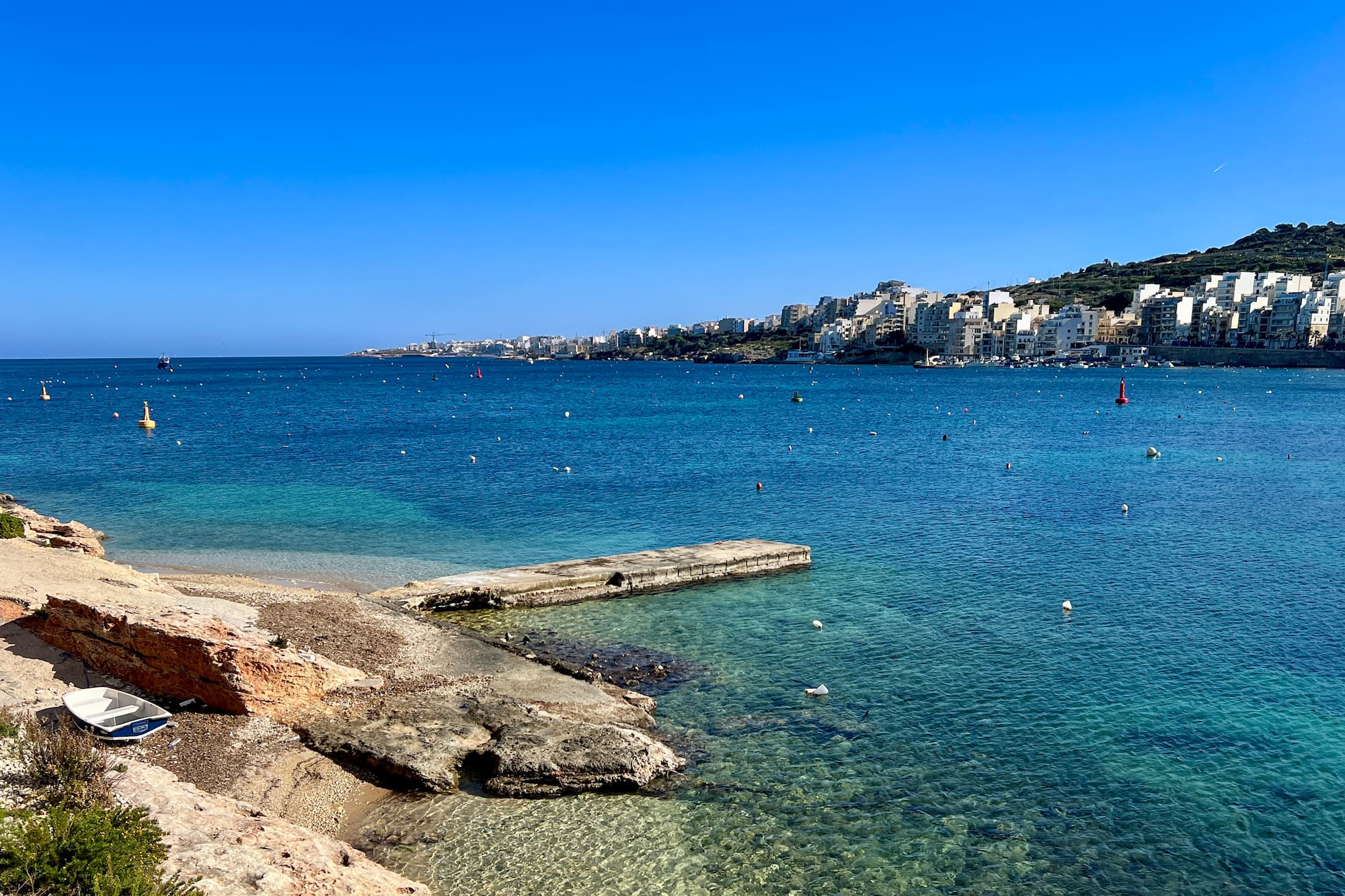 Malta: Bahía azul con pueblo costero, muelle y playa rocosa con bote.