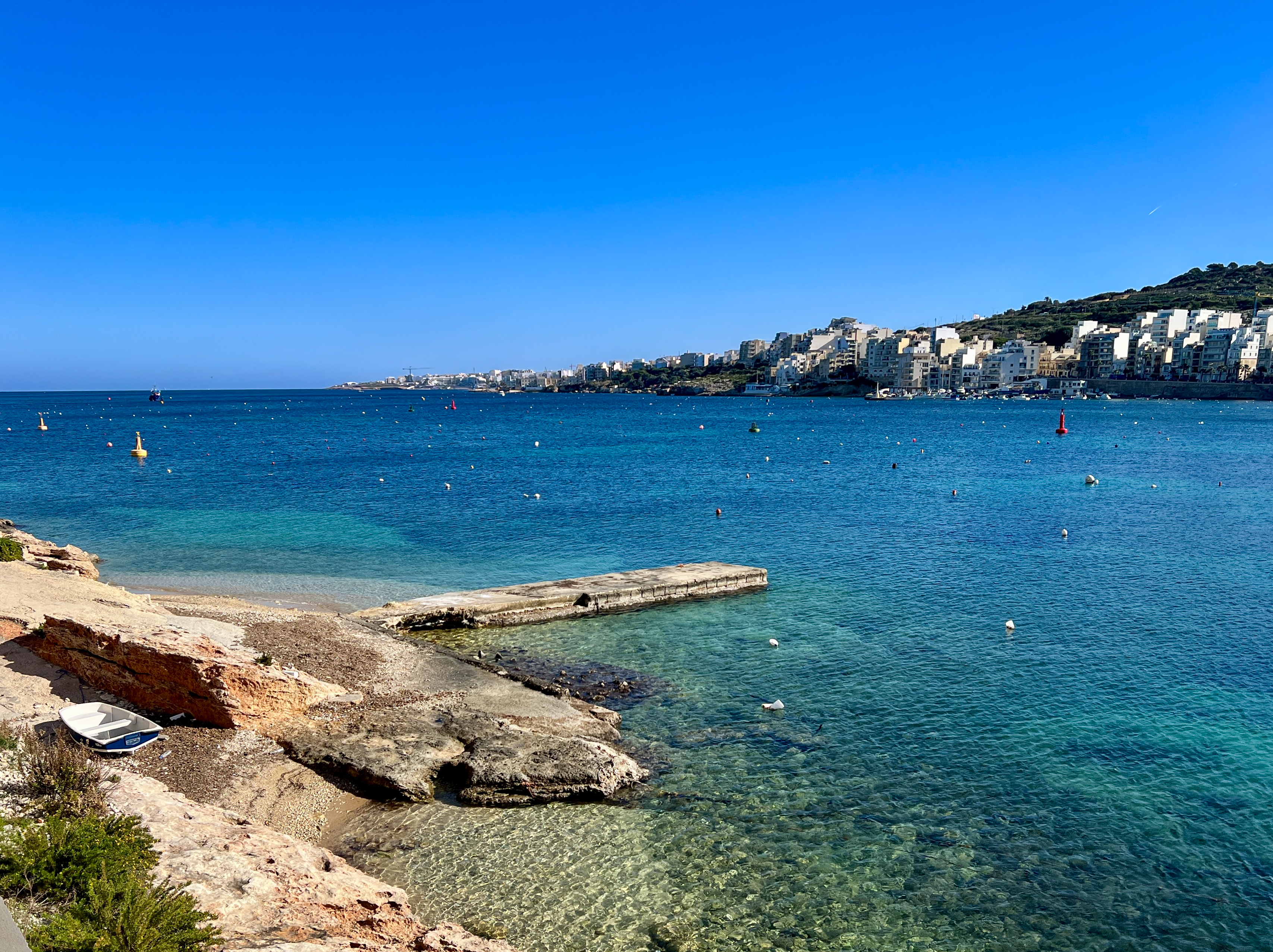 Malta: Bahía azul con pueblo costero, muelle y playa rocosa con bote.