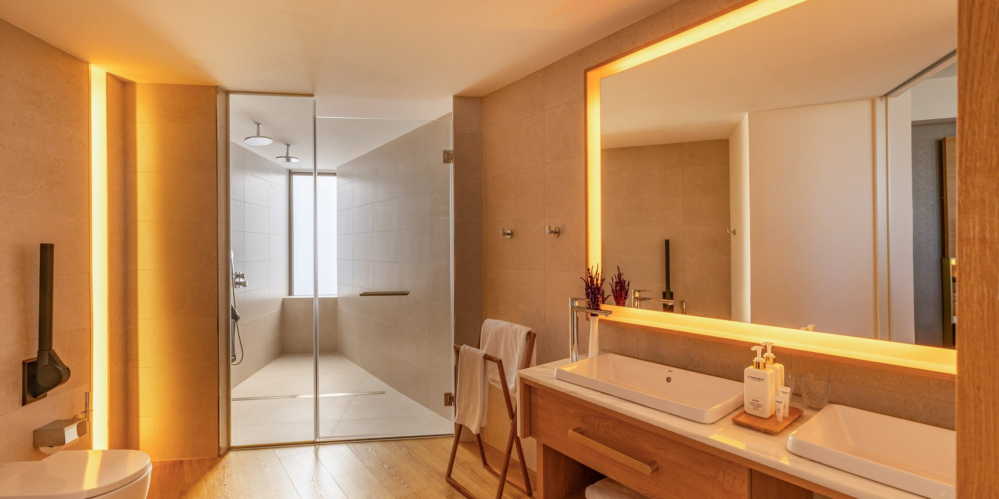 Warmly lit modern bathroom with wooden floors, glass shower, and double sink vanity.