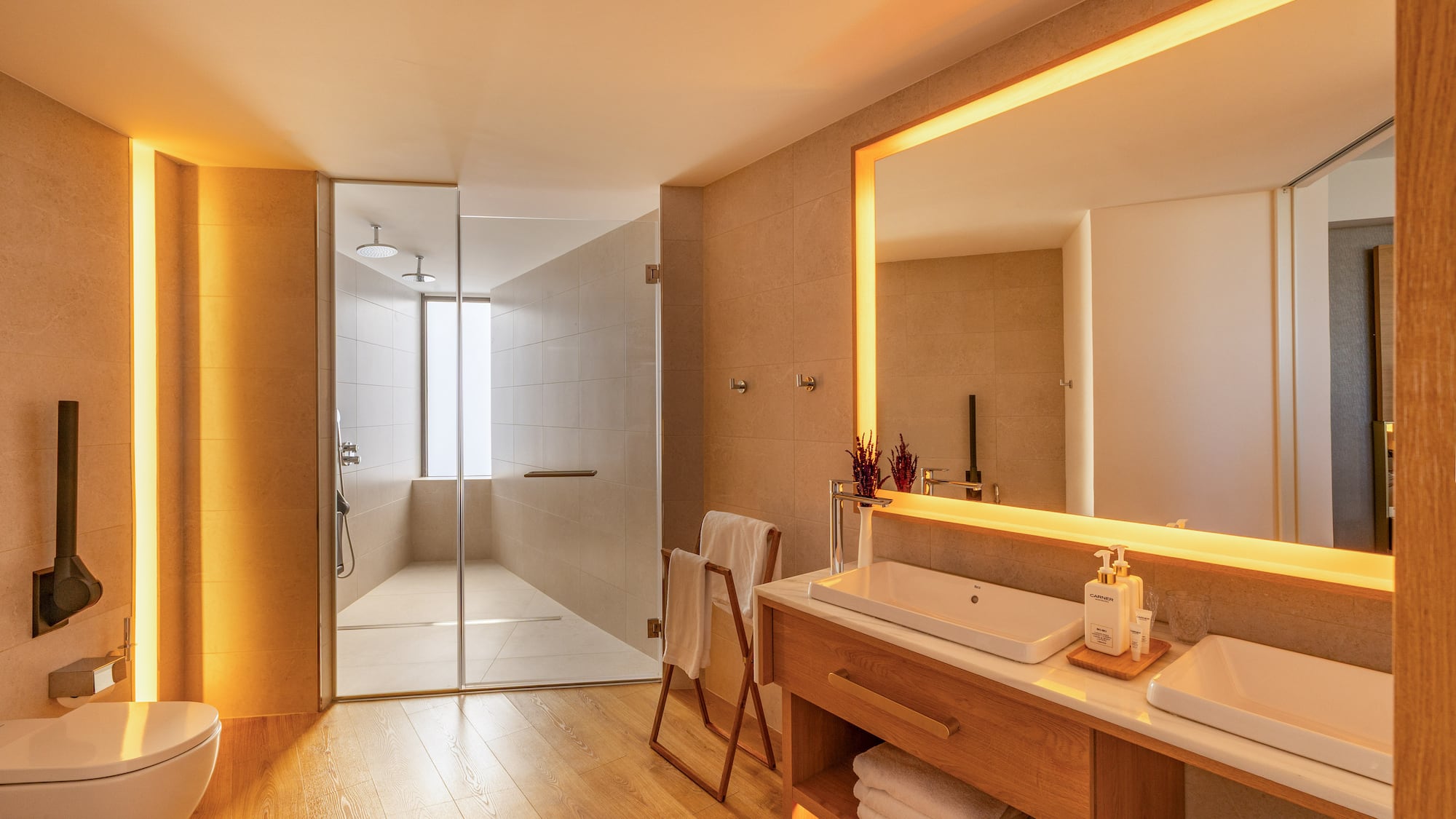 Warmly lit modern bathroom with wooden floors, glass shower, and double sink vanity.