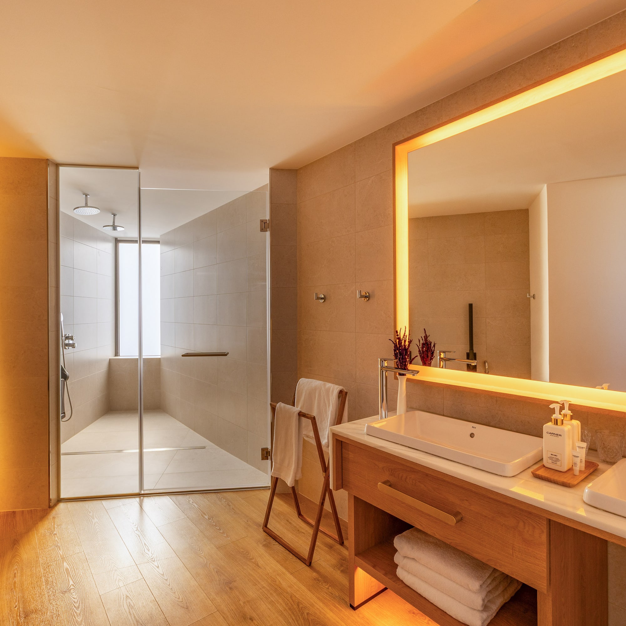 Warmly lit modern bathroom with wooden floors, glass shower, and double sink vanity.