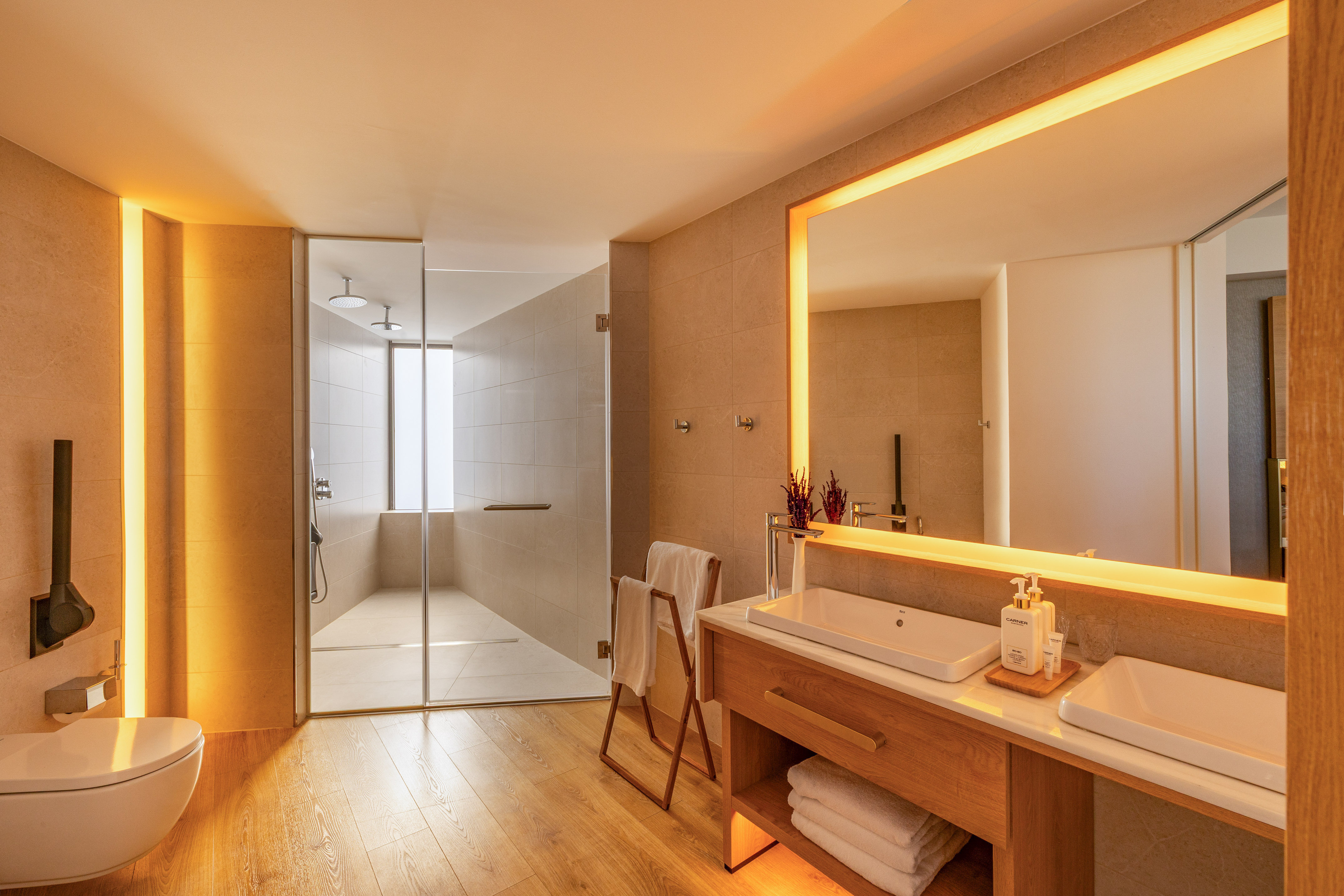 Warmly lit modern bathroom with wooden floors, glass shower, and double sink vanity.