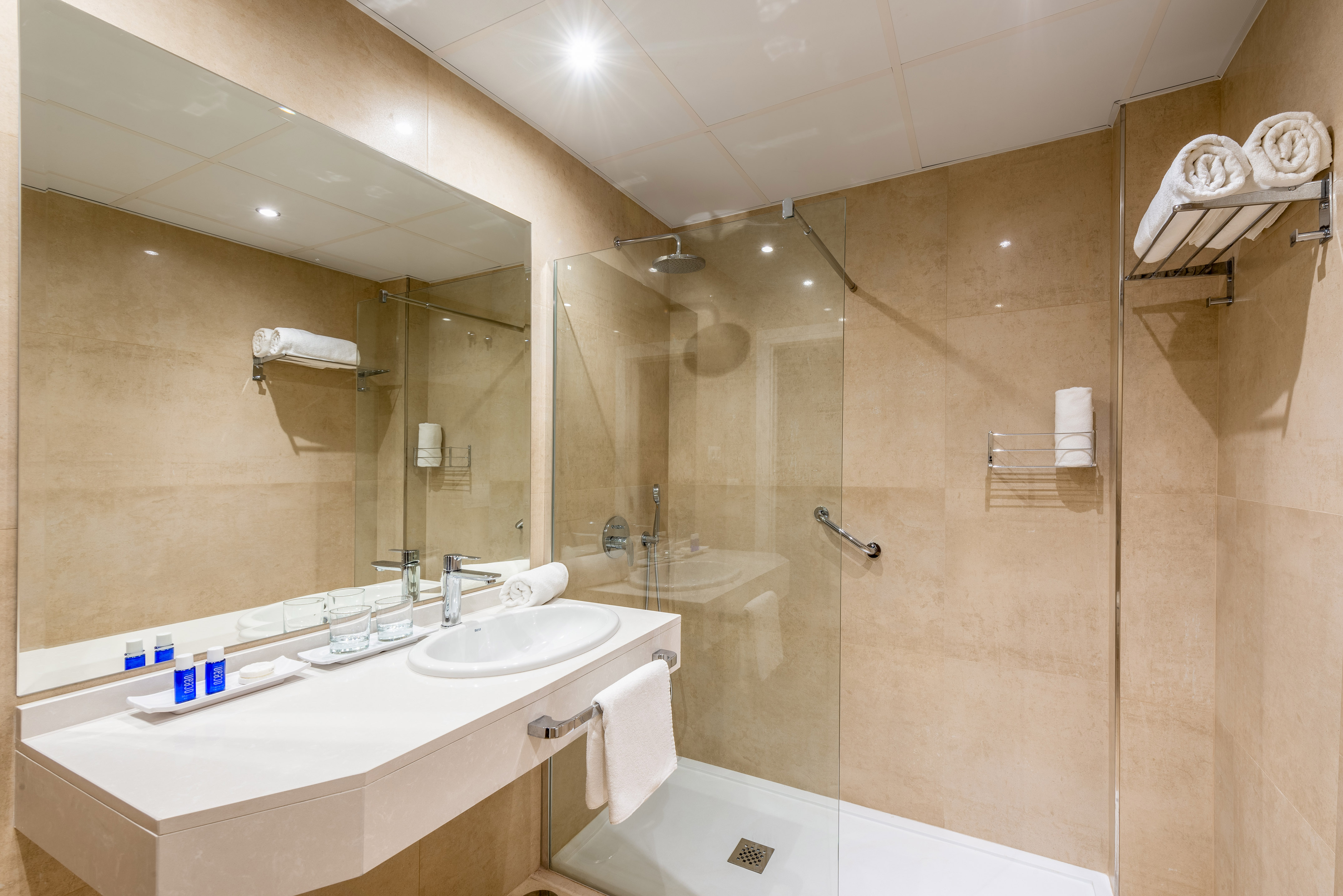 Modern hotel bathroom with beige tiles, vanity, mirror, and glass shower.