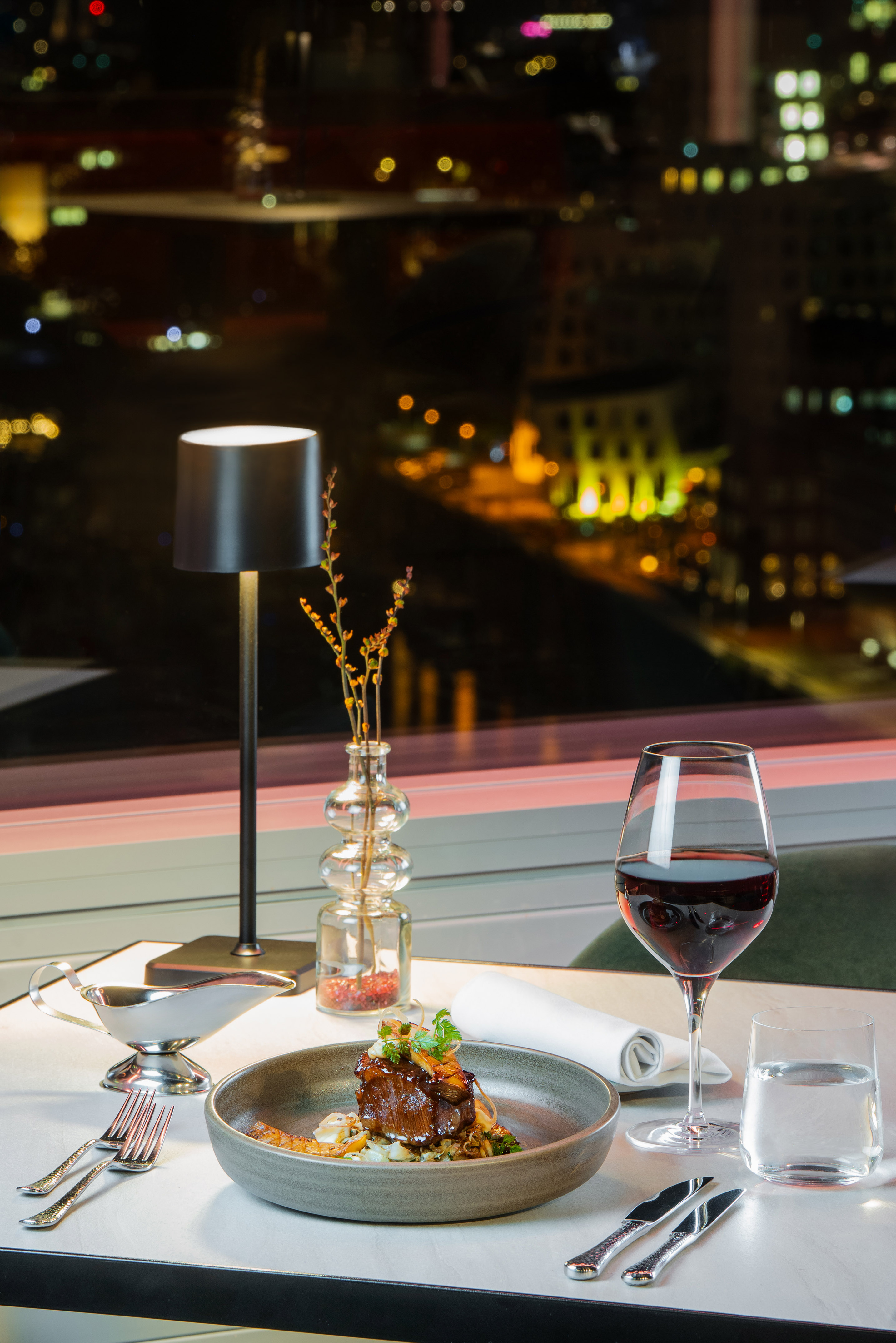 Plated gourmet meal, red wine, window view of city lights at night.