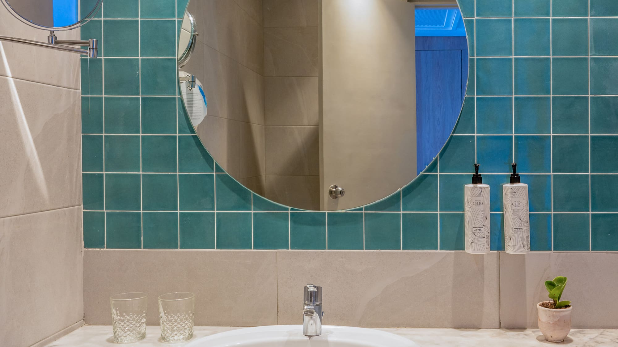 Hotel bathroom with marble vanity, white sink, round mirror, and teal wall tiles.