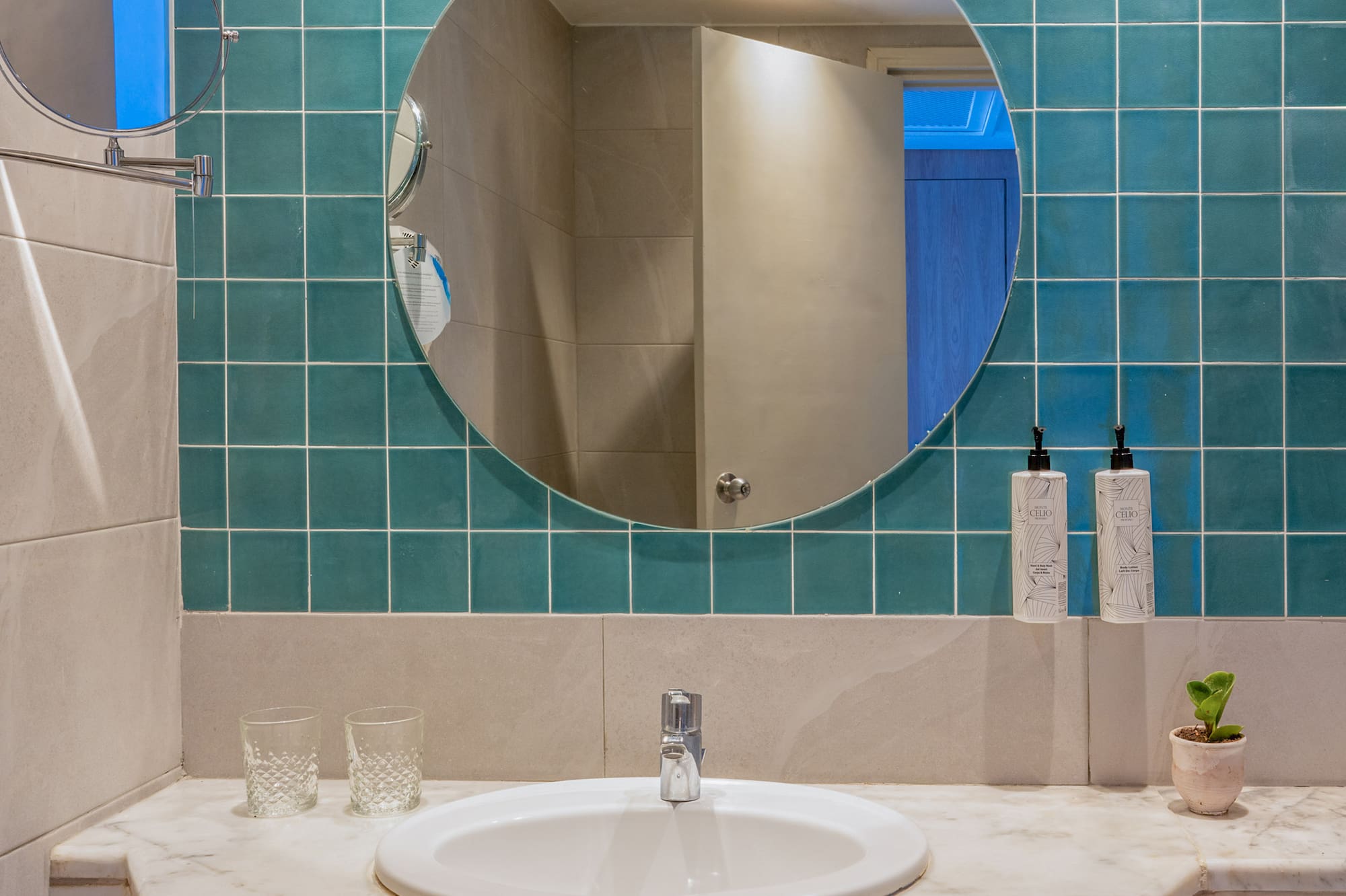 Hotel bathroom with marble vanity, white sink, round mirror, and teal wall tiles.