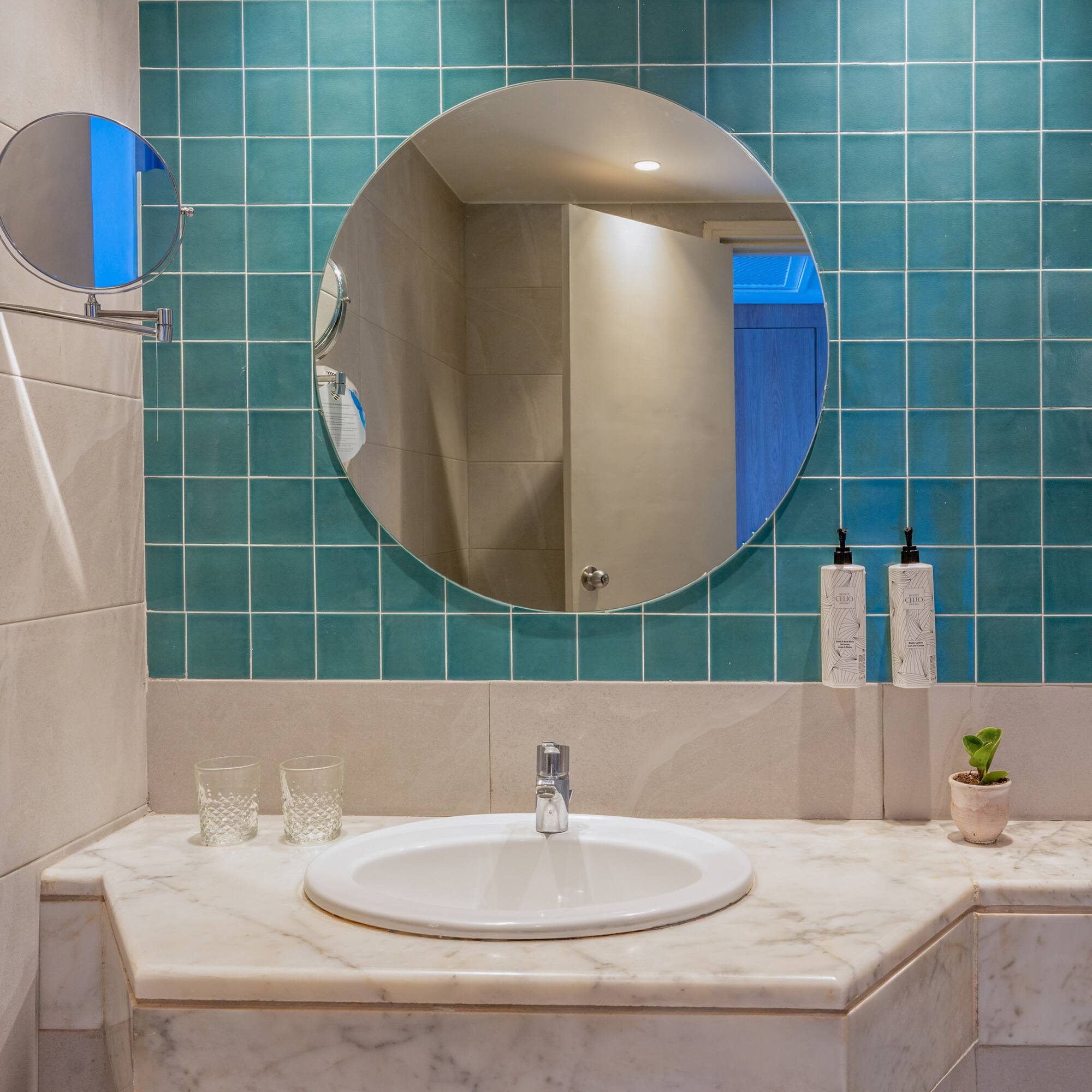 Hotel bathroom with marble vanity, white sink, round mirror, and teal wall tiles.
