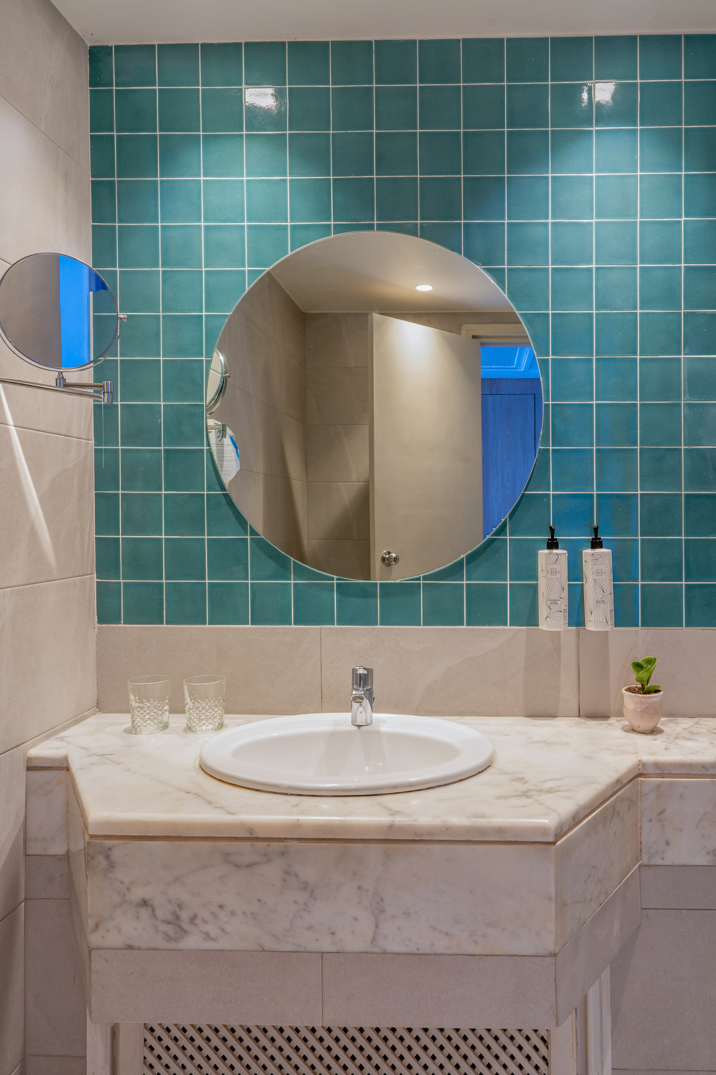 Hotel bathroom with marble vanity, white sink, round mirror, and teal wall tiles.