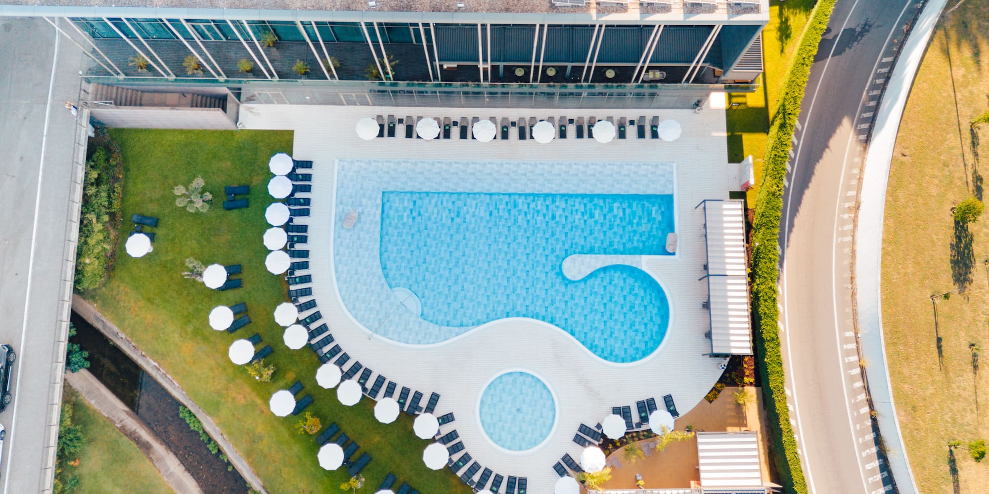 Aerial: Modern hotel pool with sun loungers, umbrellas, green lawns.