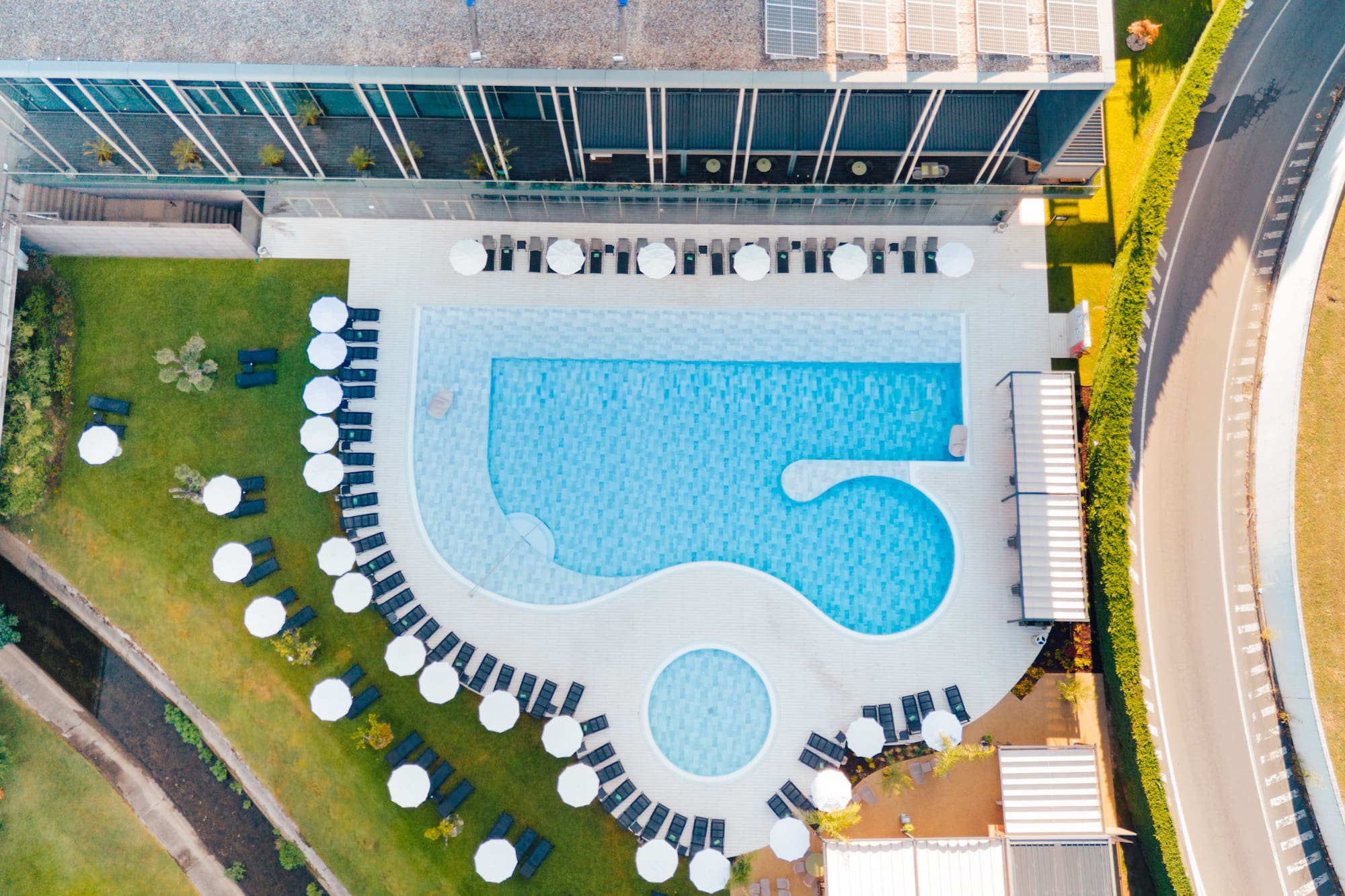 Aerial: Modern hotel pool with sun loungers, umbrellas, green lawns.