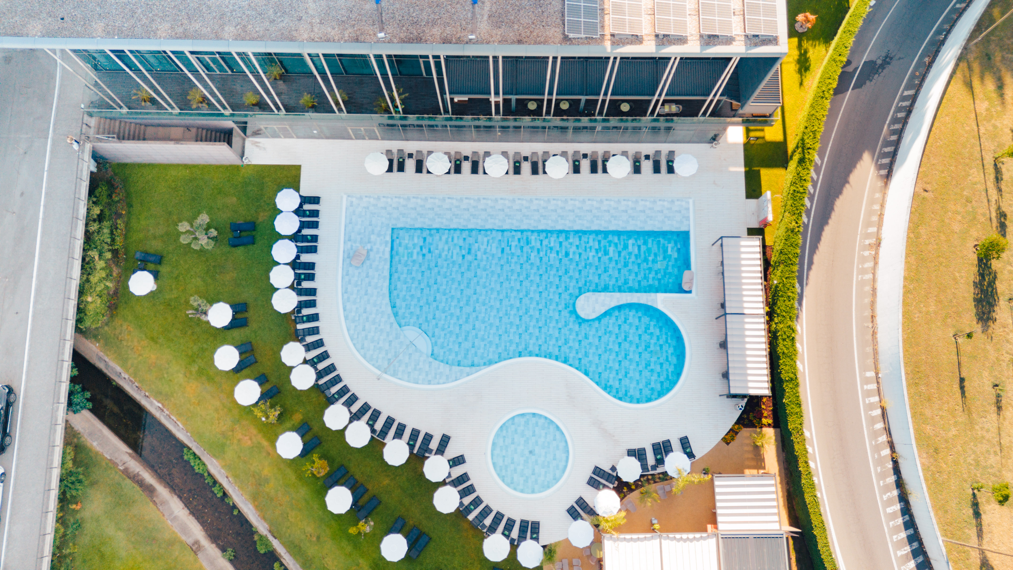 Aerial: Modern hotel pool with sun loungers, umbrellas, green lawns.